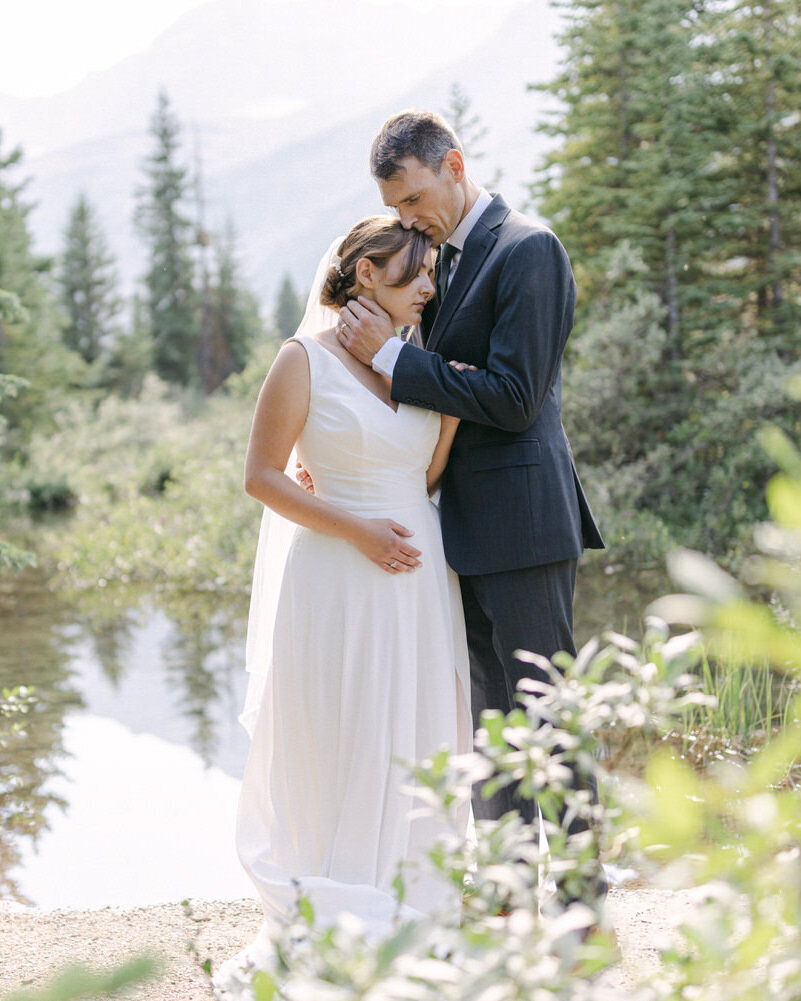 This serene image captures the bride and groom holding each other as they walk through a forested area near Bow Lake. The intimate embrace and the natural beauty of the forest leading to the expansive waters of Bow Lake create a picturesque and romantic setting, reflecting the deep connection between the couple.