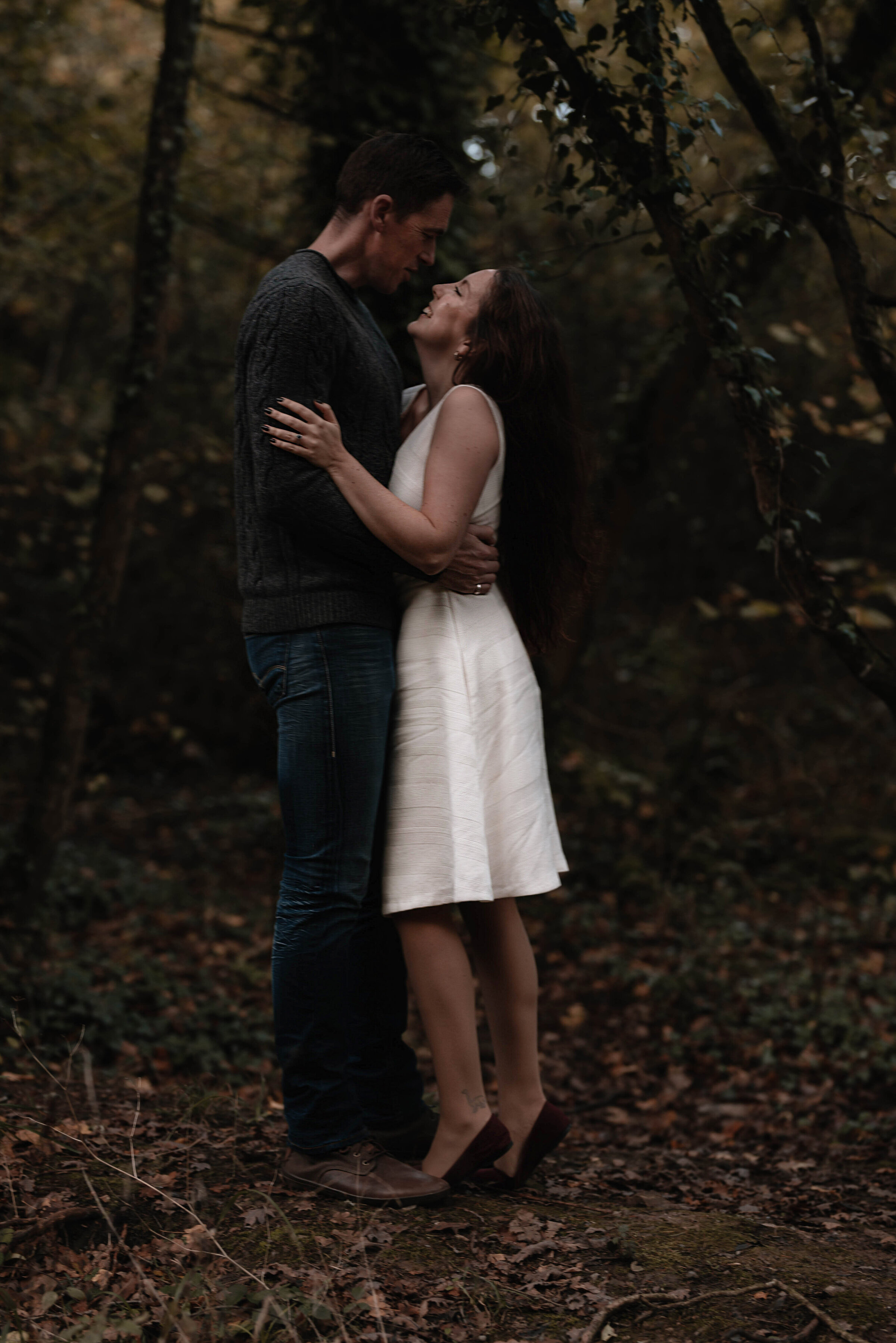 Woman looking up at man and smiling whilst they embrace in a wooded area surrounded by trees and fallen leaves