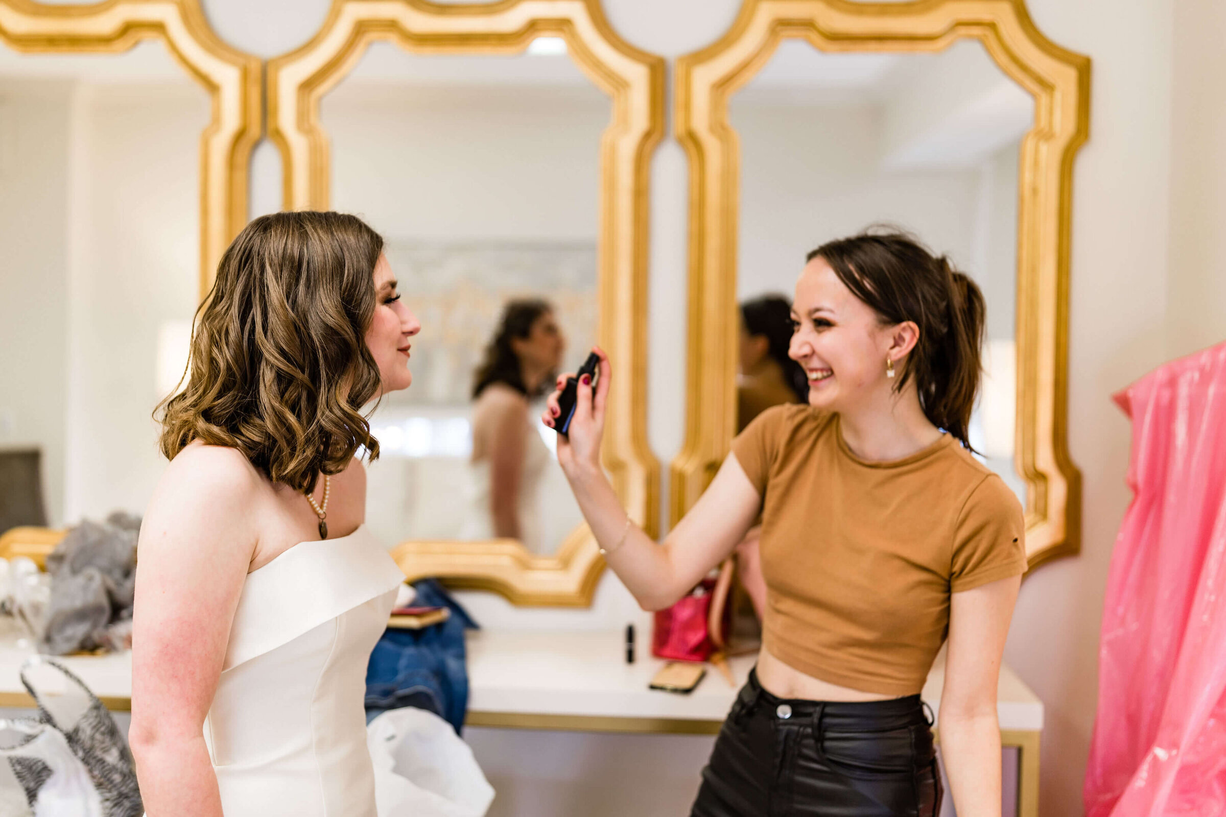 A smiling makeup artist wearing a brown crop top and black pants applies makeup to a woman in a white strapless dress in front of two gold-framed mirrors.