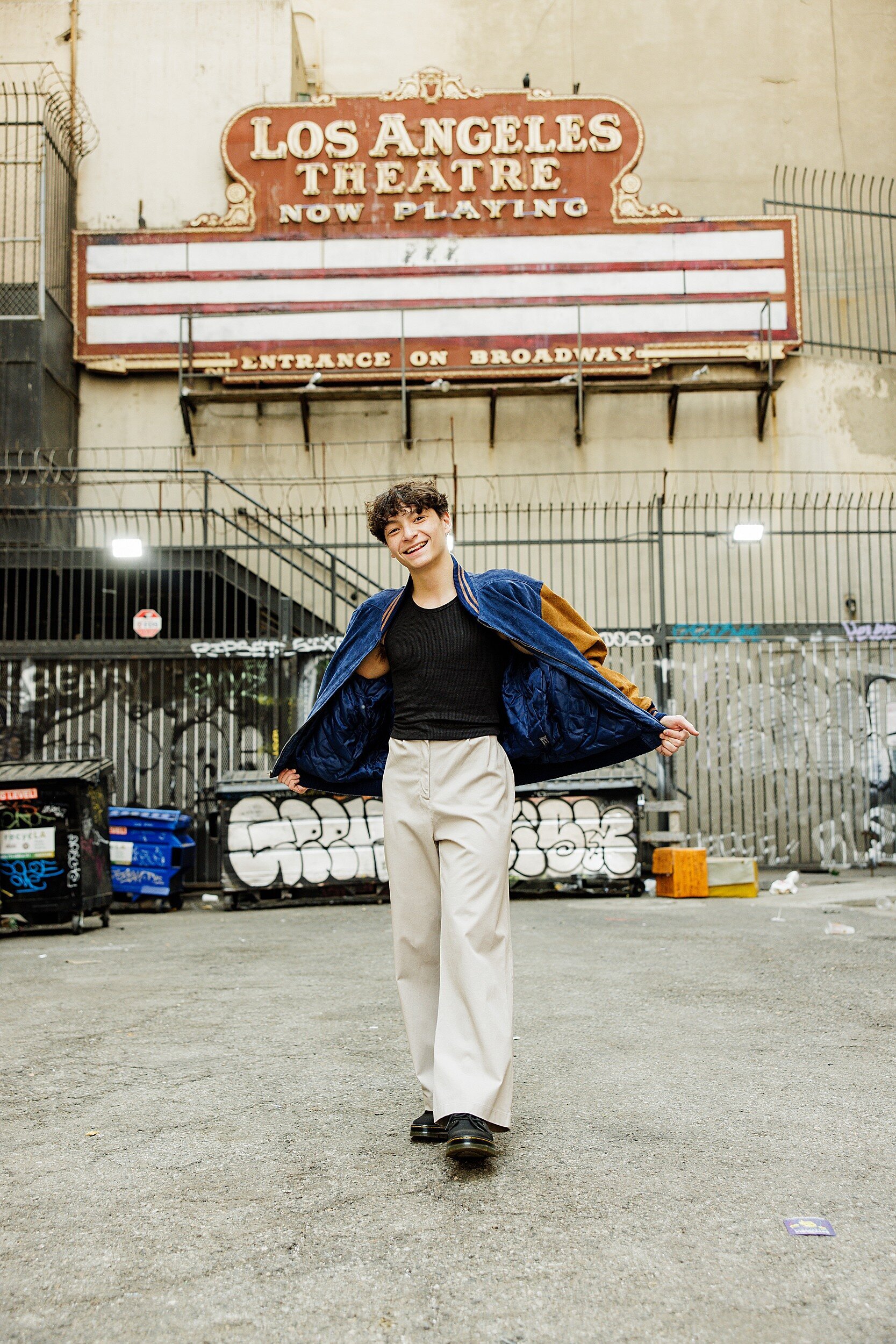 Fashion-forward senior guy in cream trousers and a black tee flashes his jacket open while smiling in front of graffiti walls and an old theater marquee in Downtown Los Angeles.