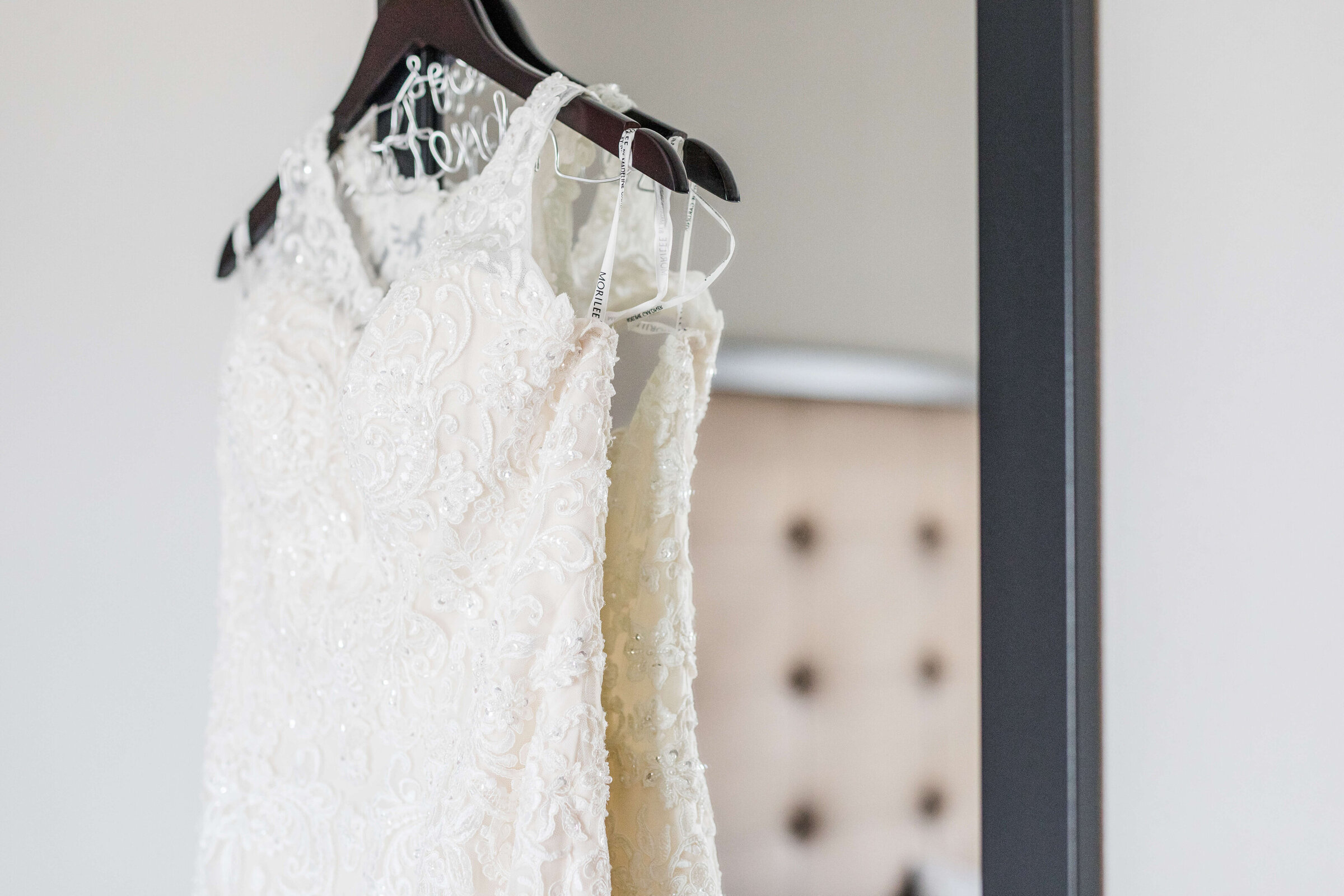 A wedding gown hanging on a mirror in a hotel room. You can see the reflection of the bed in the mirror.