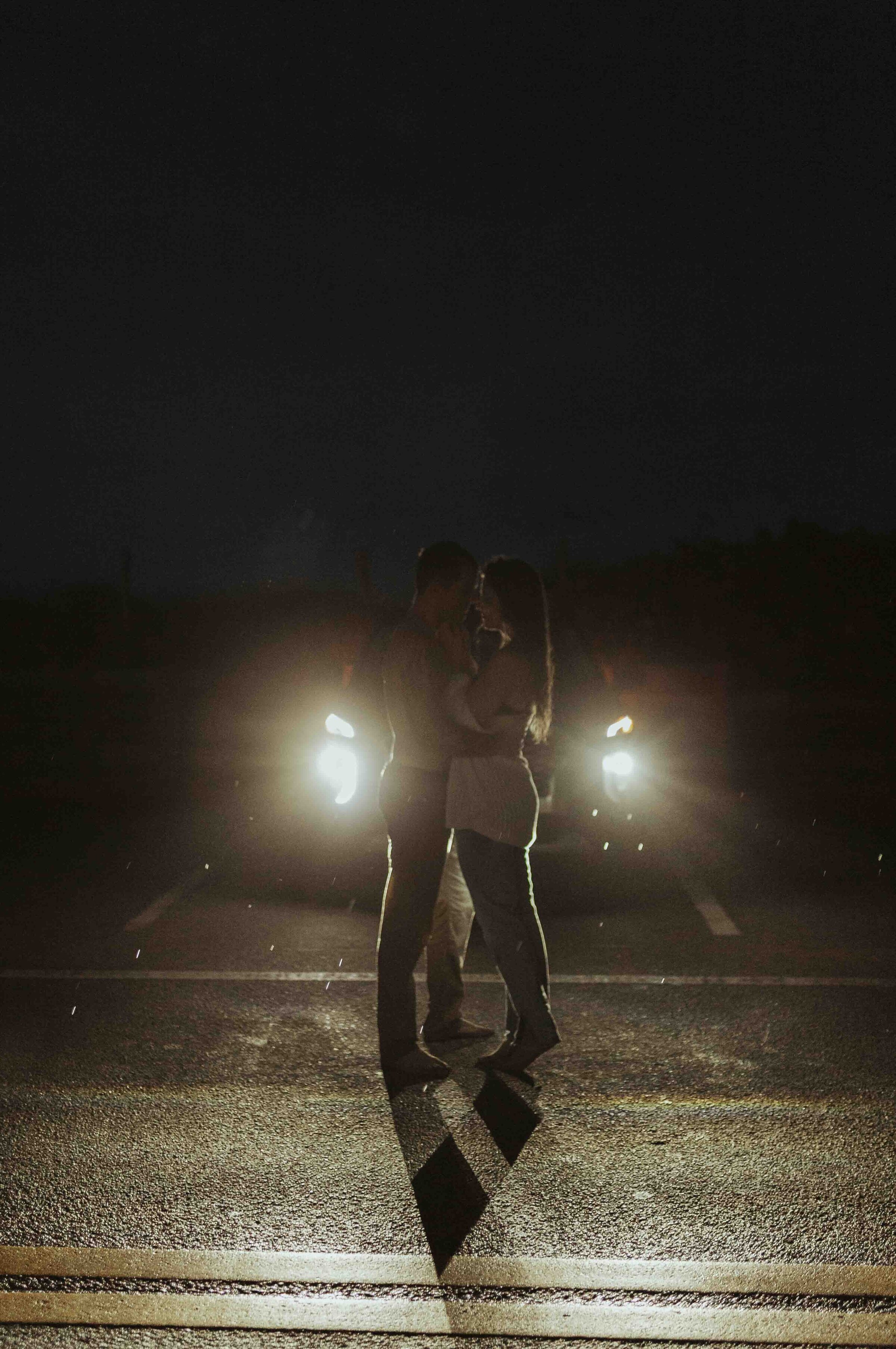 Couple embracing in a parking lot at night with car headlines