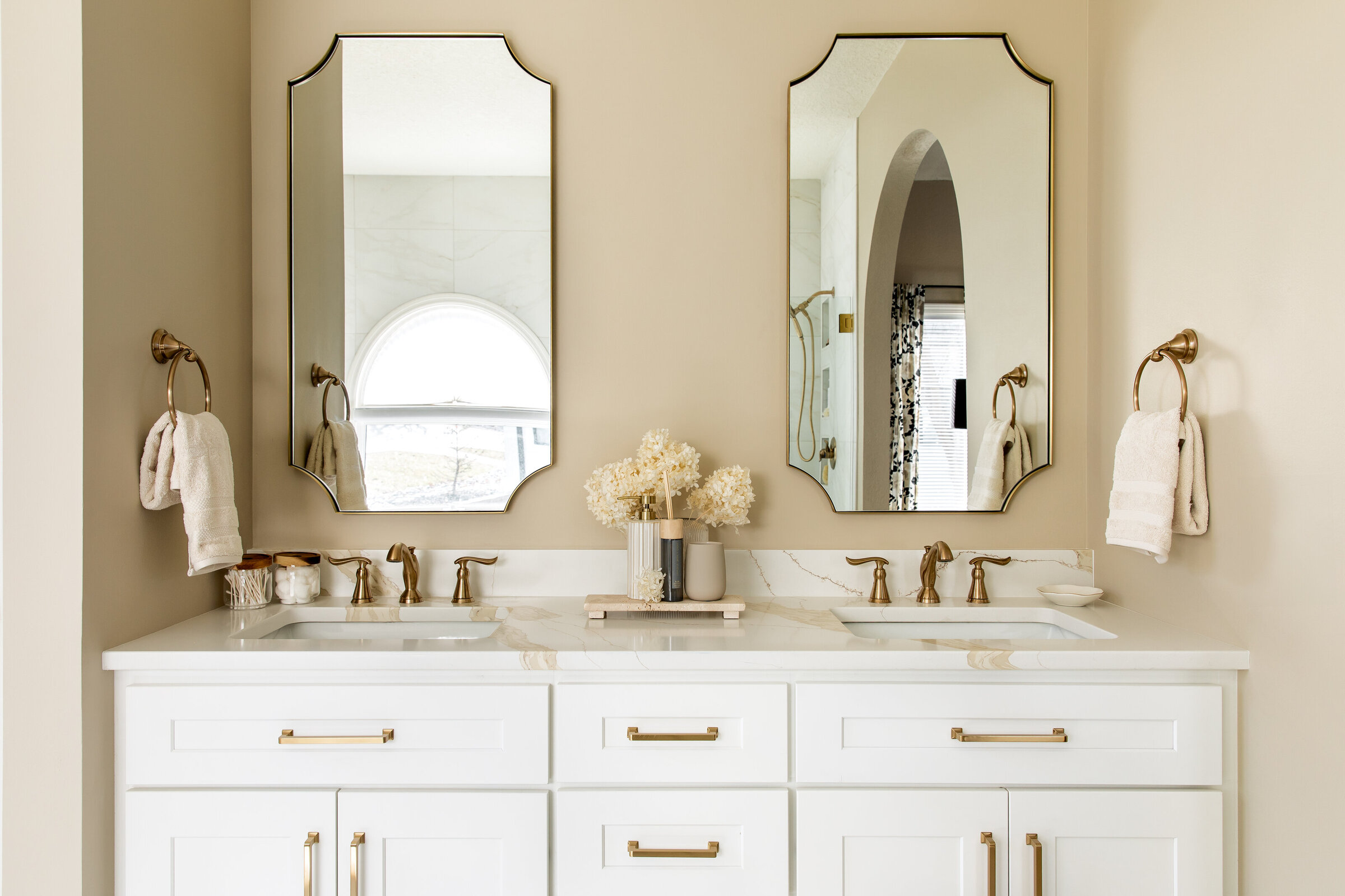 Large white double sink vanity with gold mirrors, hardware and plumbing fixtures featuring a neutral beige wall paint.