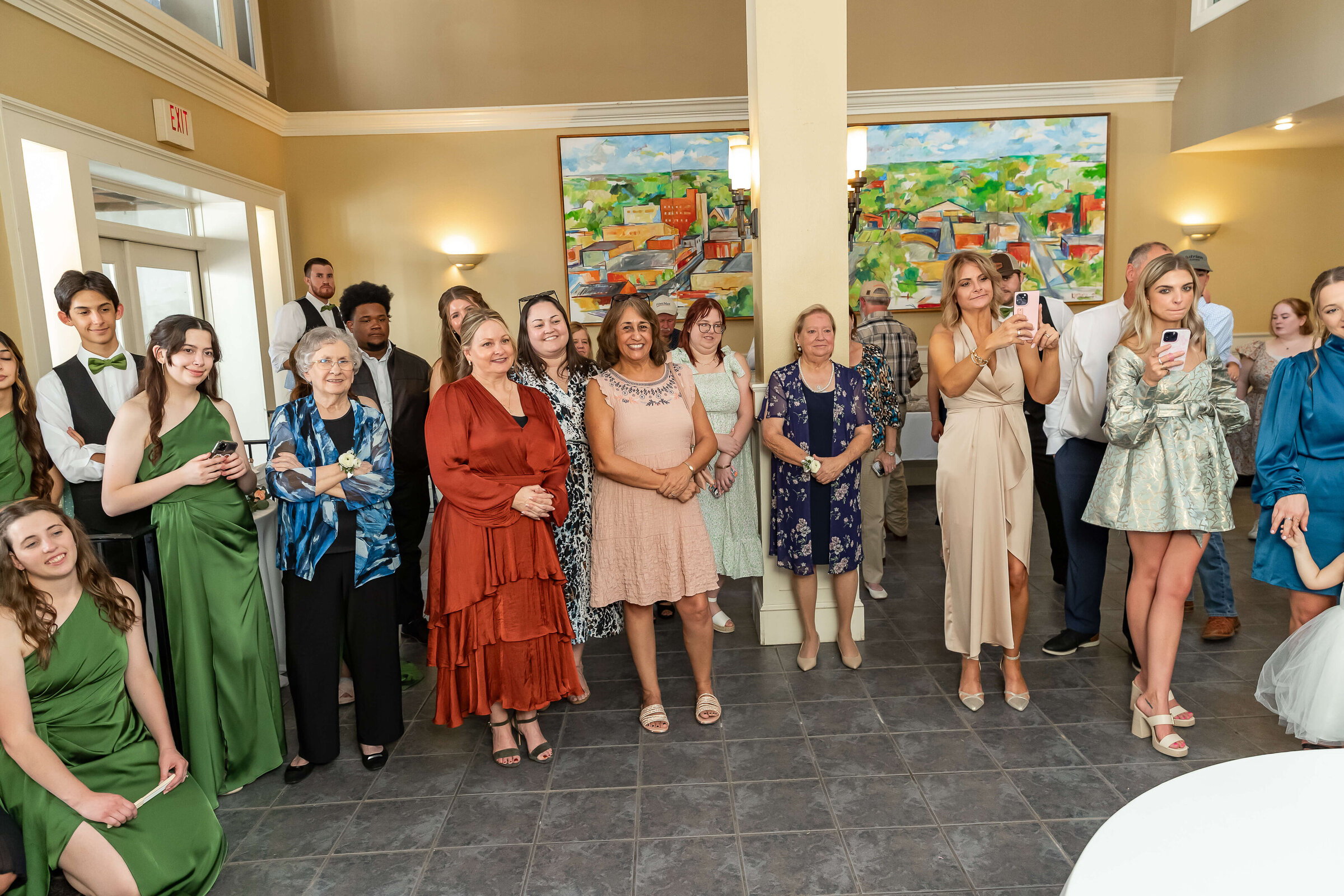 A group of people dressed in semi-formal attire stand indoors, smiling and watching something off-camera; some are taking photos with phones. The room is bright.