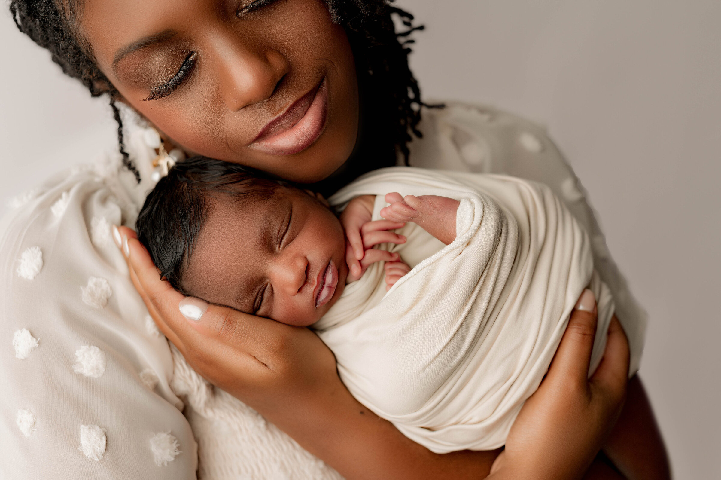 A woman lovingly cradles a sleeping newborn wrapped in a cream blanket. As a Charlotte newborn photographer takes her photo, she gently holds the baby close to her face, smiling softly. The background is blurred, highlighting this tender moment captured perfectly in North Carolina.