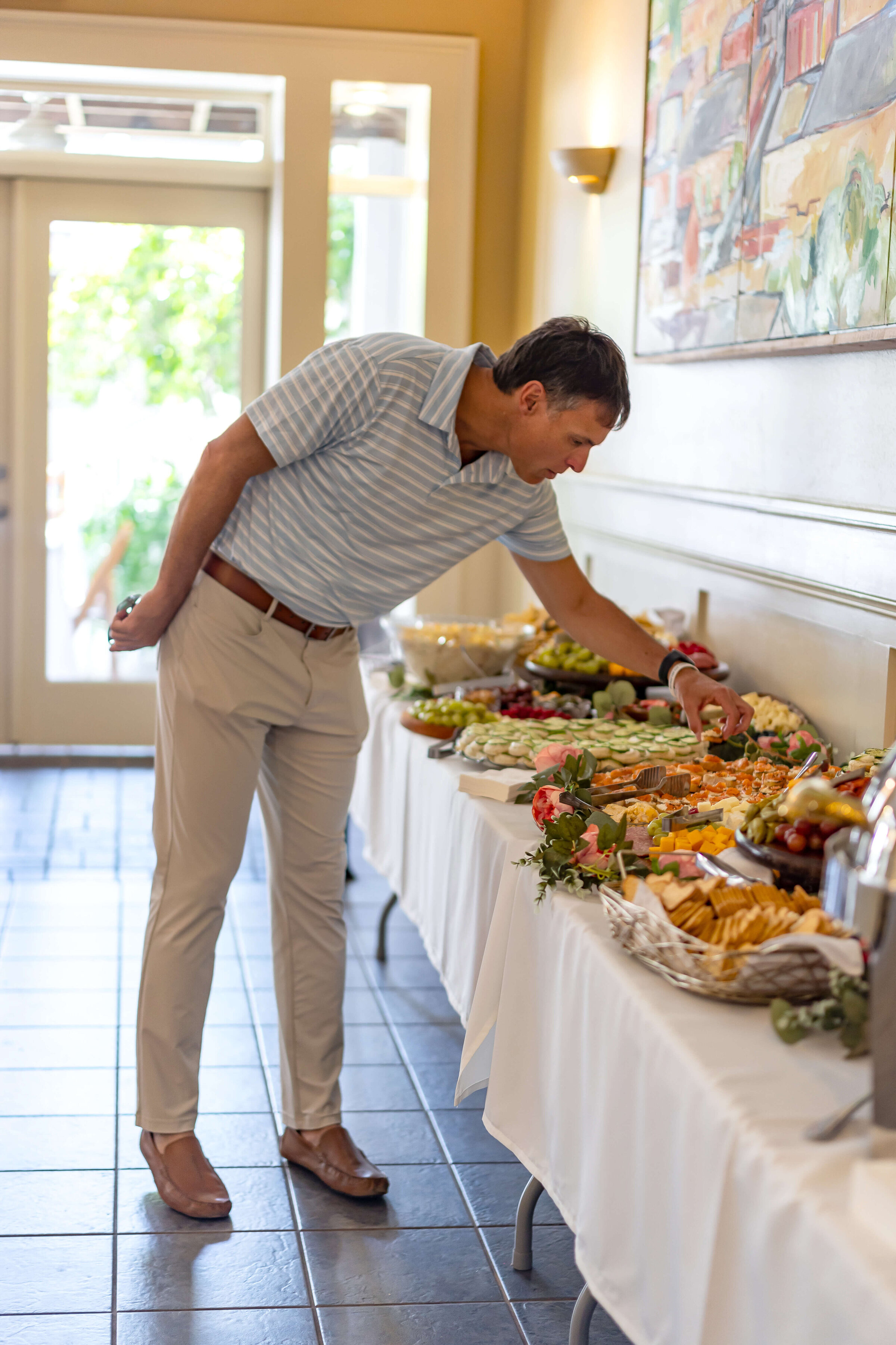 A man in a light blue striped shirt and beige pants is selecting food from a buffet table covered with assorted dishes and flowers in a bright room.