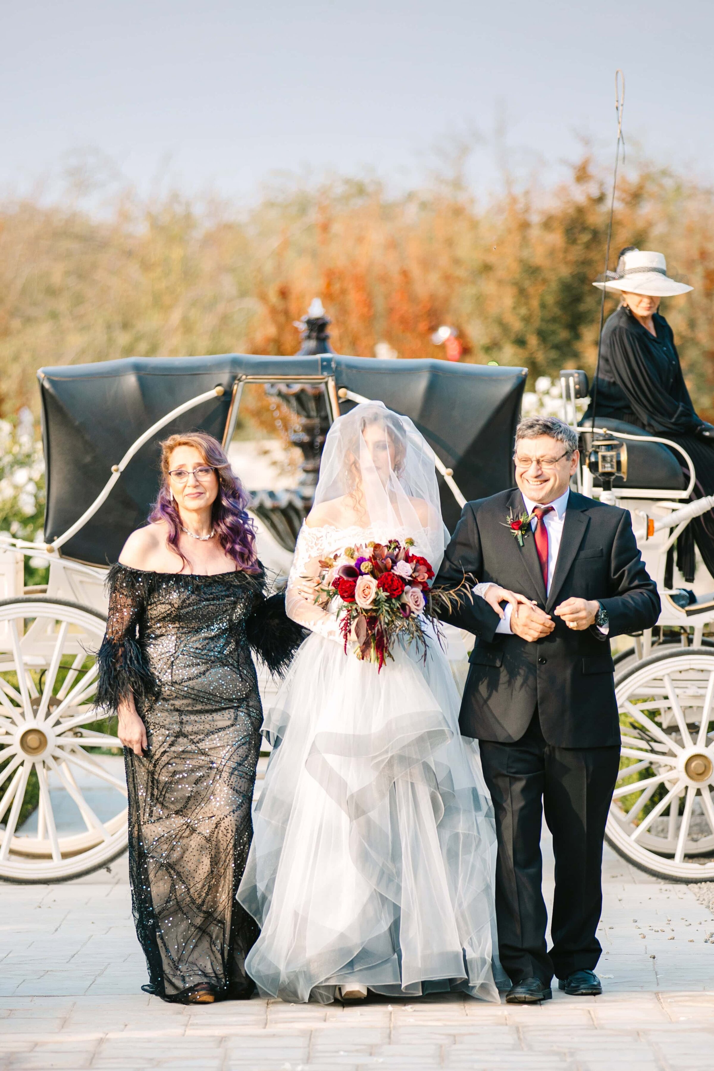 Bride wakling down the wedding aisle with both her mother and father with a horse drawn carriage behind them