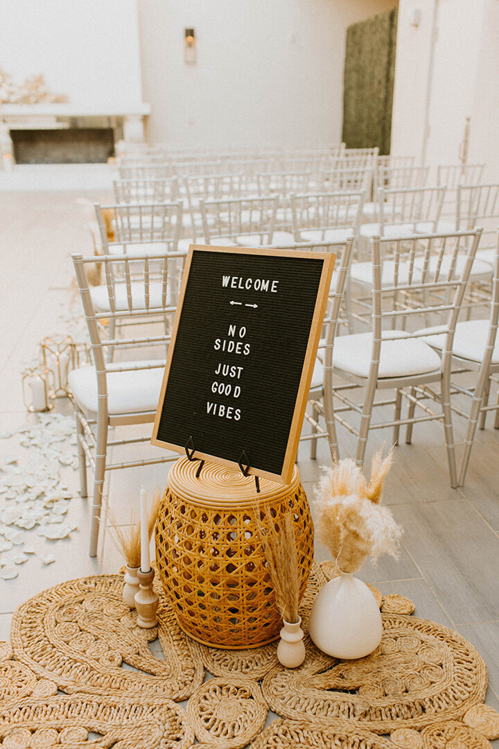 A decorative sign reading “Welcome — No Sides, Just Good Vibes” stands at the entrance of a ceremony setup at SoHo63, a Chandler wedding and event venue. Surrounded by pampas grass, candles, and rattan accents, the aisle is lined with silver chairs and white rose petals, showcasing the venue’s chic, modern-boho wedding style.