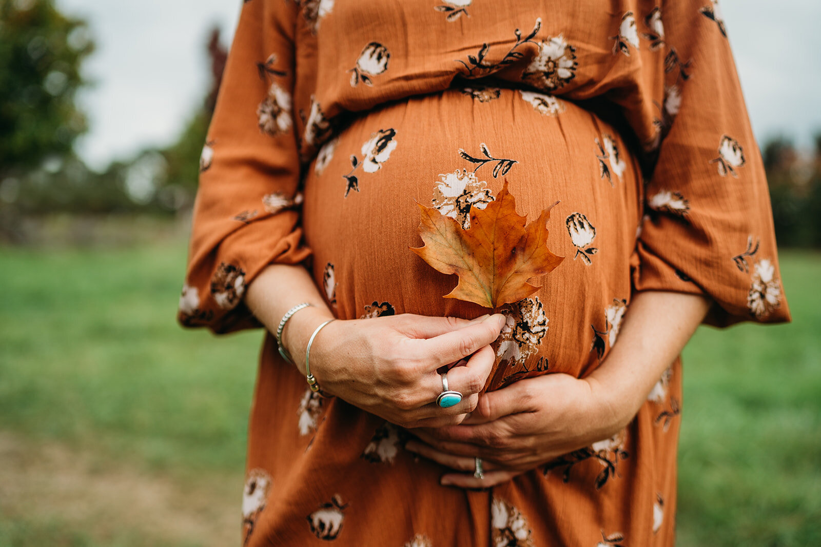 woman in burnt orange maternity dress holds autumn leaf to belly
