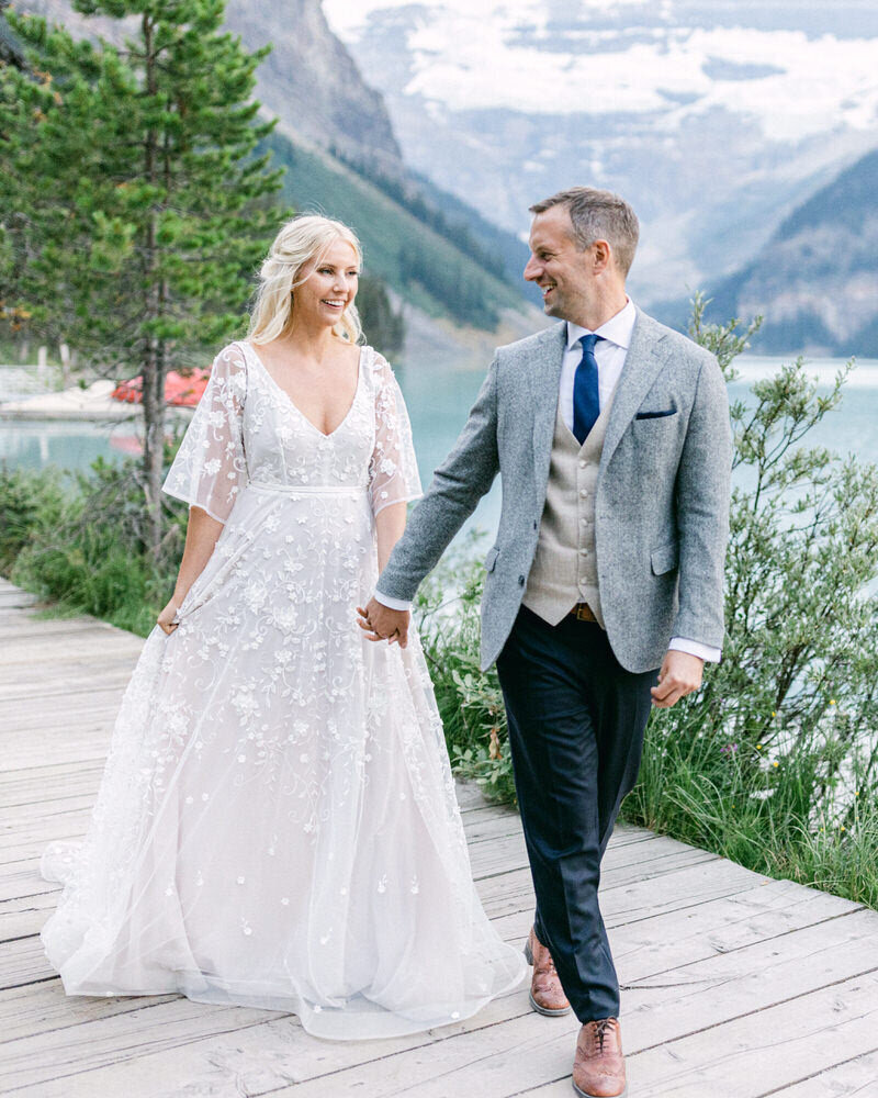 This joyful image captures the bride and groom walking hand in hand around Lake Louise, passing the iconic boat house. Their happiness shines through as they enjoy this picturesque moment, surrounded by the stunning natural beauty of the lake and mountains.