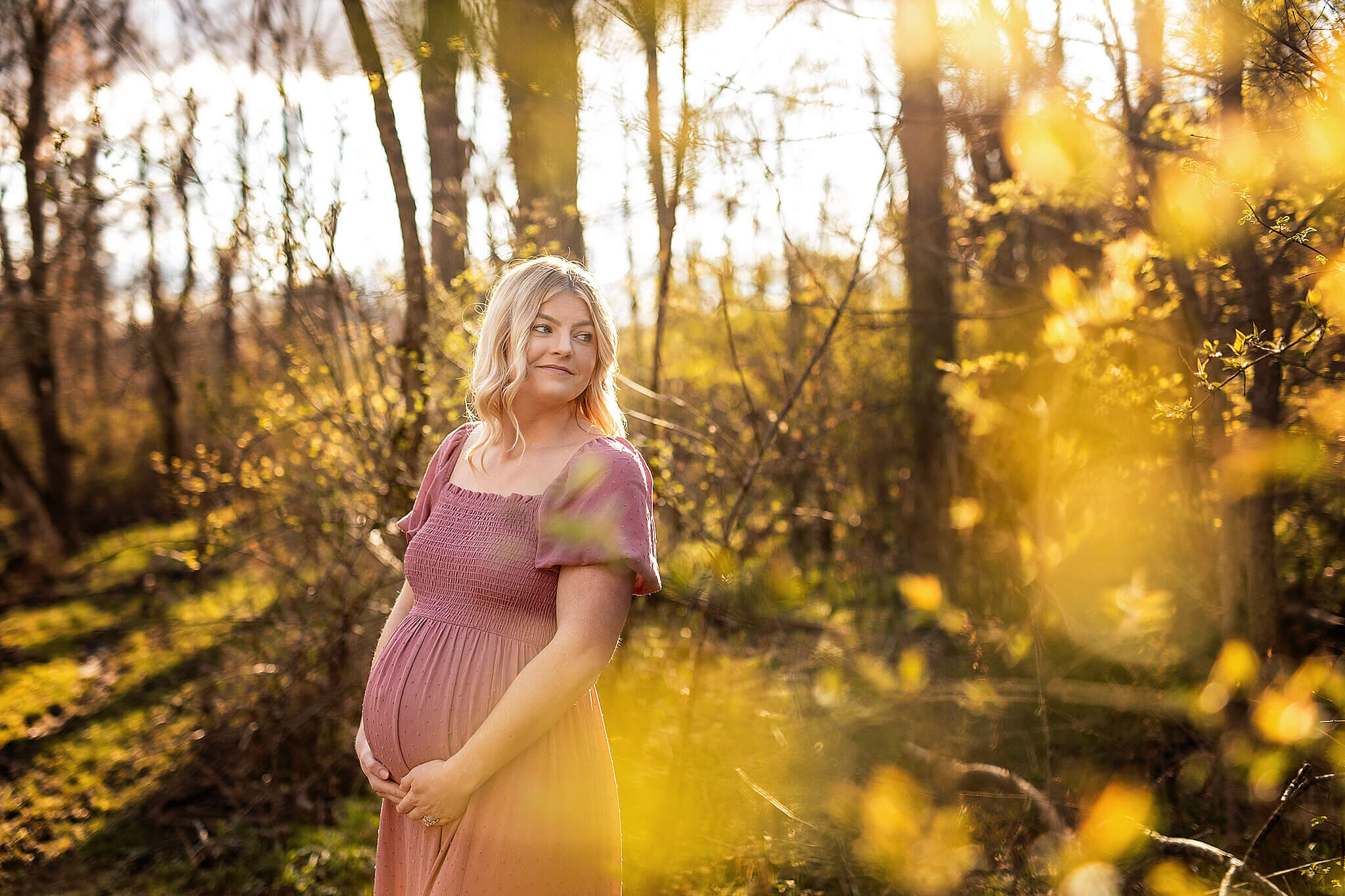 Pregnant women in pink dress looking behind her shoulder while holding her belly, standing in the woods with pretty light behind her