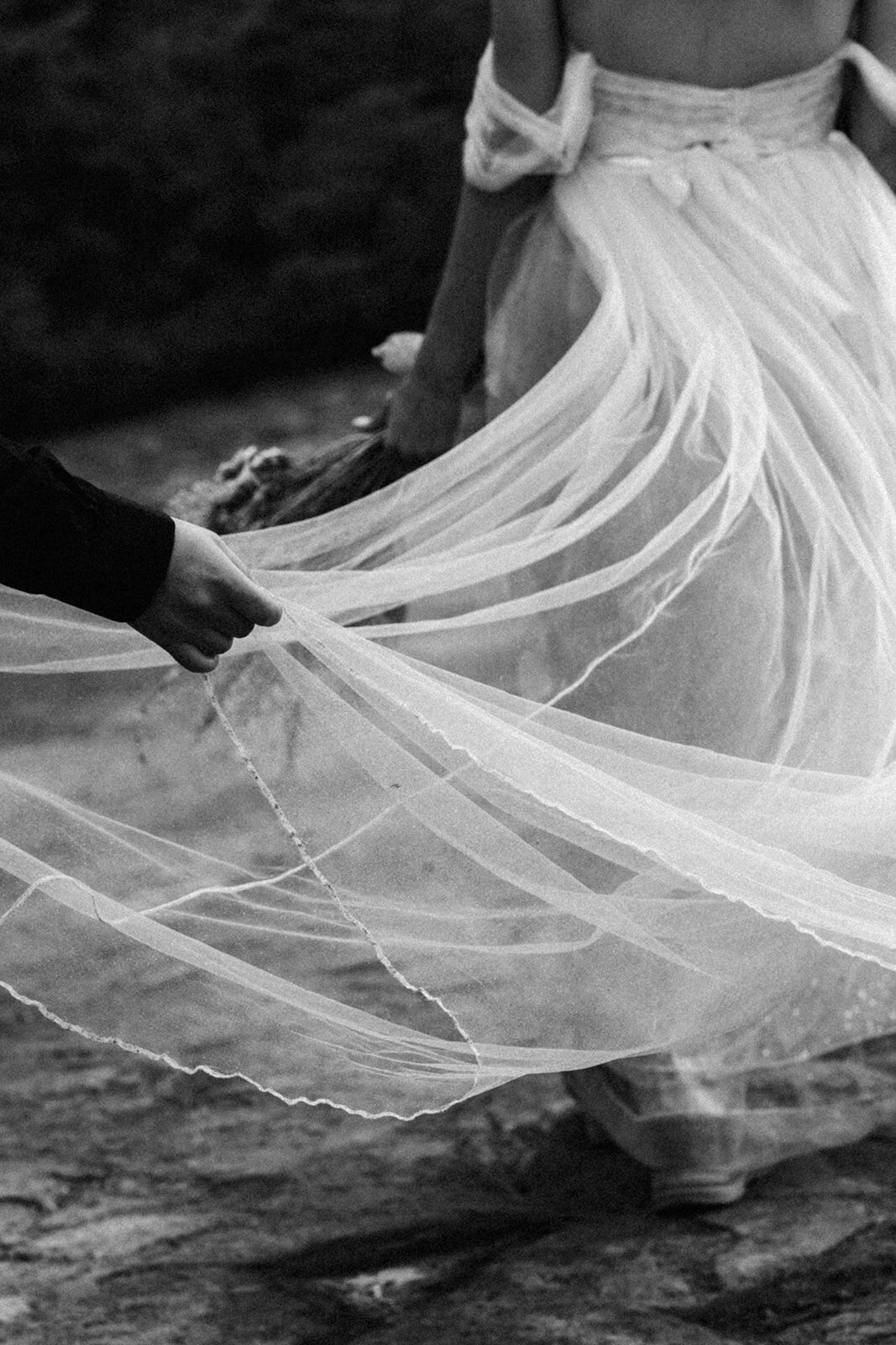 A black and white image of a groom holding the train of his brides chiffon wedding dress train. this tightly cropped image highlights the movement of the fabric from the billowing winds. 