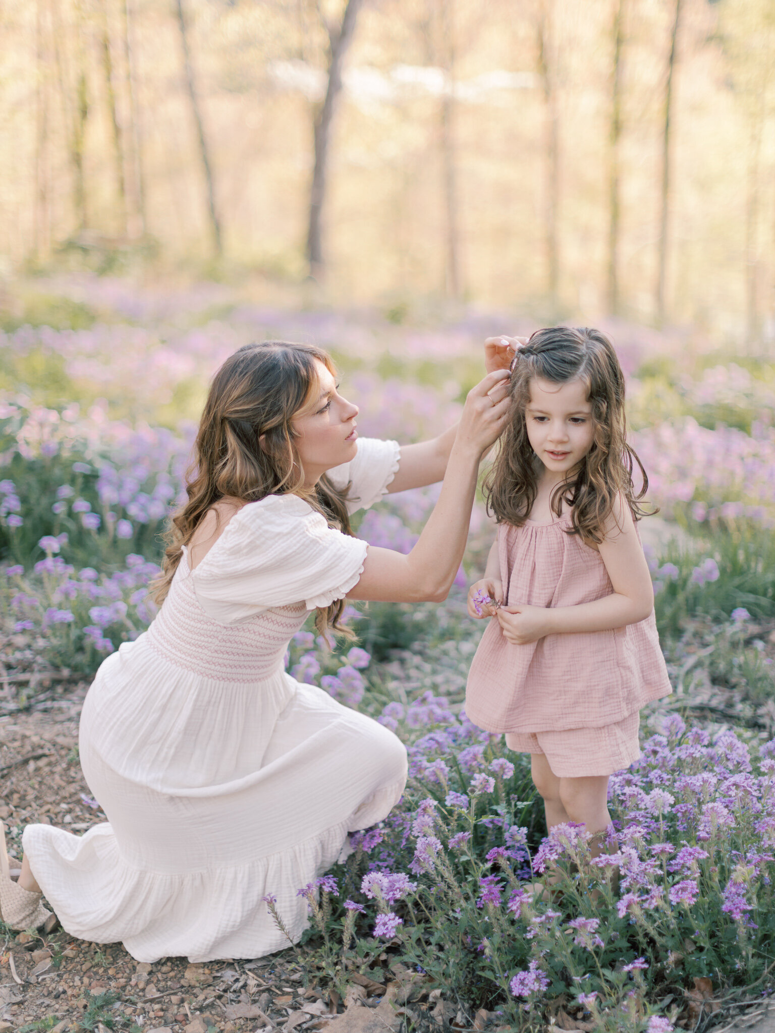 Bailey Feeler, Little Rock photographer, kneels in purple flowers to fix young brunette daughter's hair