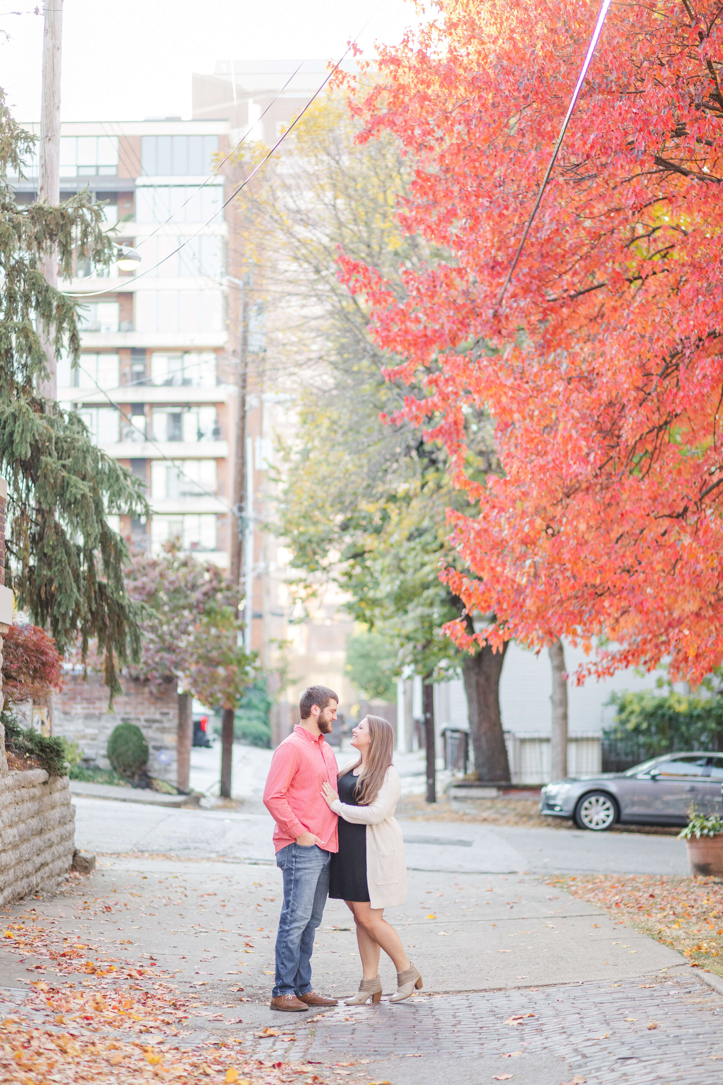 A man and woman hold each other in a downtown covington kentucky park. It's a wide shot with red fall trees in the background. She's wearing a black dress and he's in jeans and a red shirt. Taken by a kentucky wedding photographer