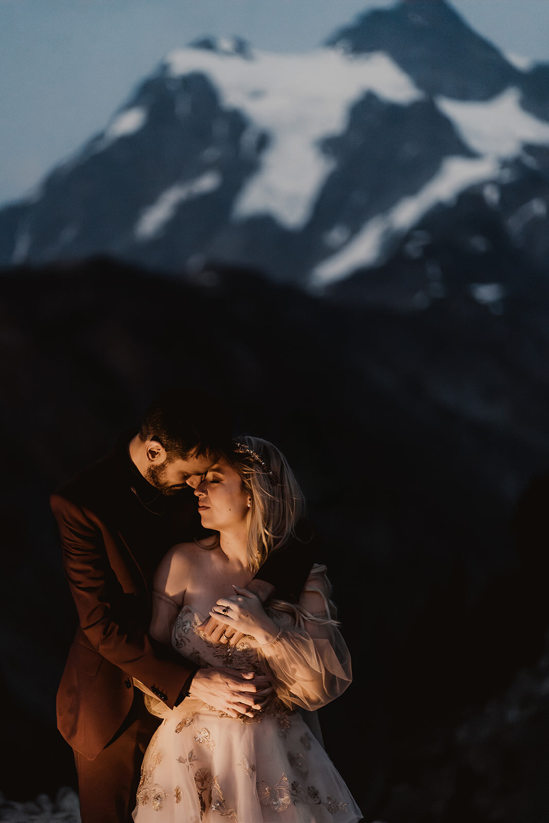 a couple in their wedding attire embrace, pressing their heads against each other. A snow-capped mountain paints their background