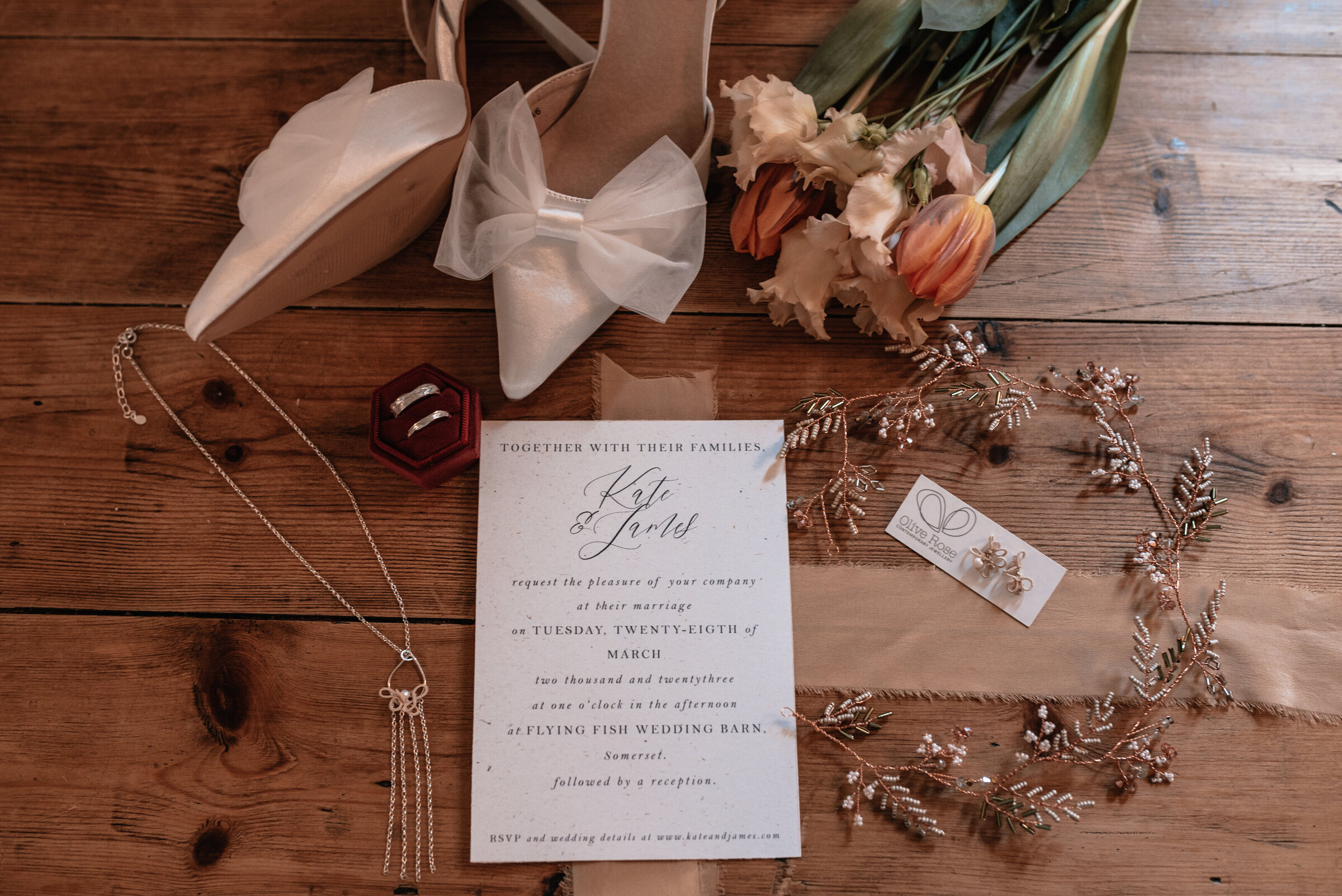 Wedding invitation surrounded by bridal jewellery, wedding rings, bridal shoes and bouquet on a wooden table