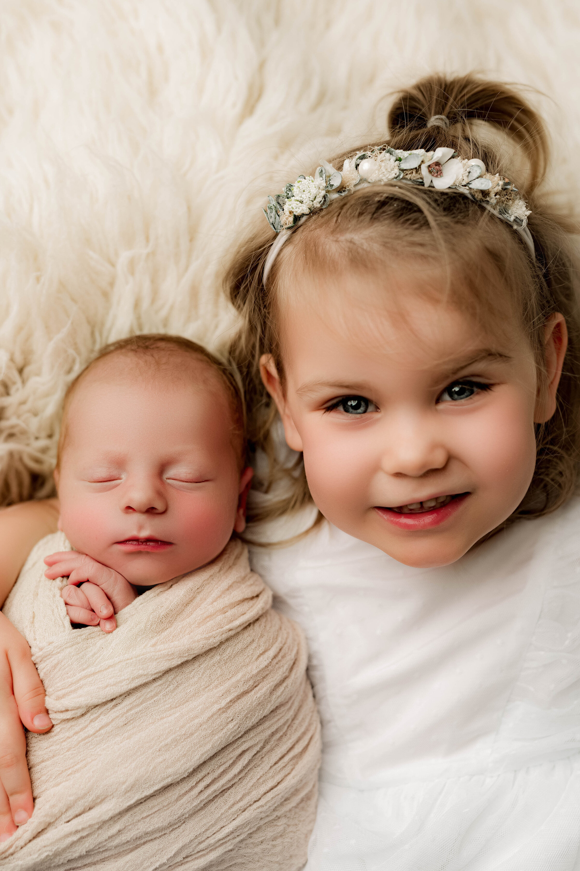 A young child with a floral headband smiles beside a sleeping baby wrapped in a beige blanket. They rest on a fluffy, cream-colored surface, creating a cozy atmosphere captured beautifully by a North Carolina newborn photographer.