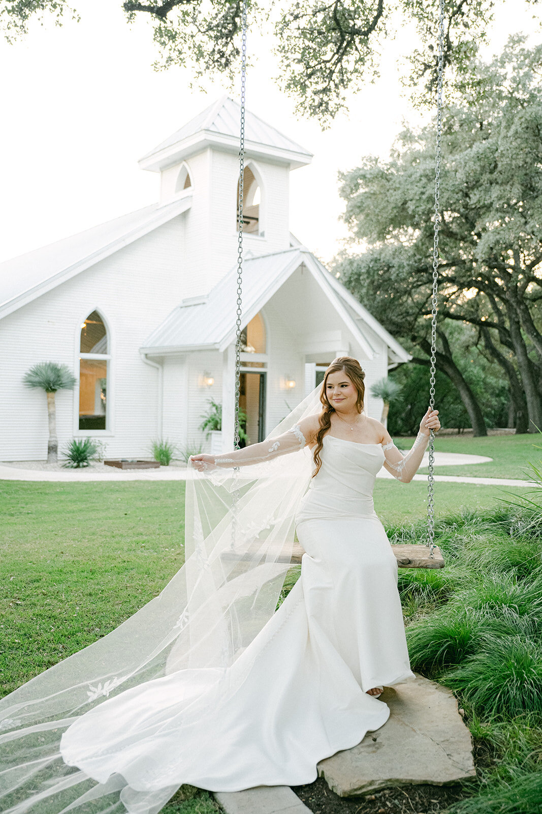 Bride on a swing outside the white chapel of The Chandelier of Gruene in Texas with a long train flowing, captured during golden hour following the ceremony.