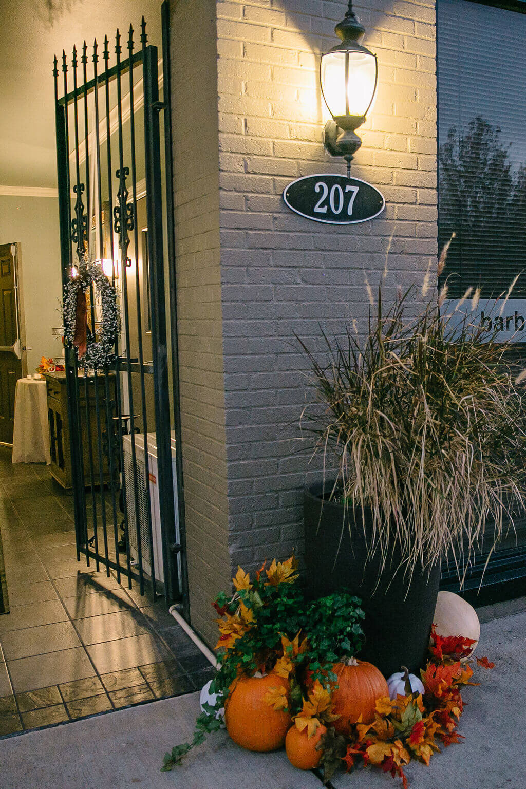 A warmly lit entryway with a black metal gate, building number 207, potted grasses, and pumpkins with autumn leaves arranged at the door, creating a festive fall atmosphere.