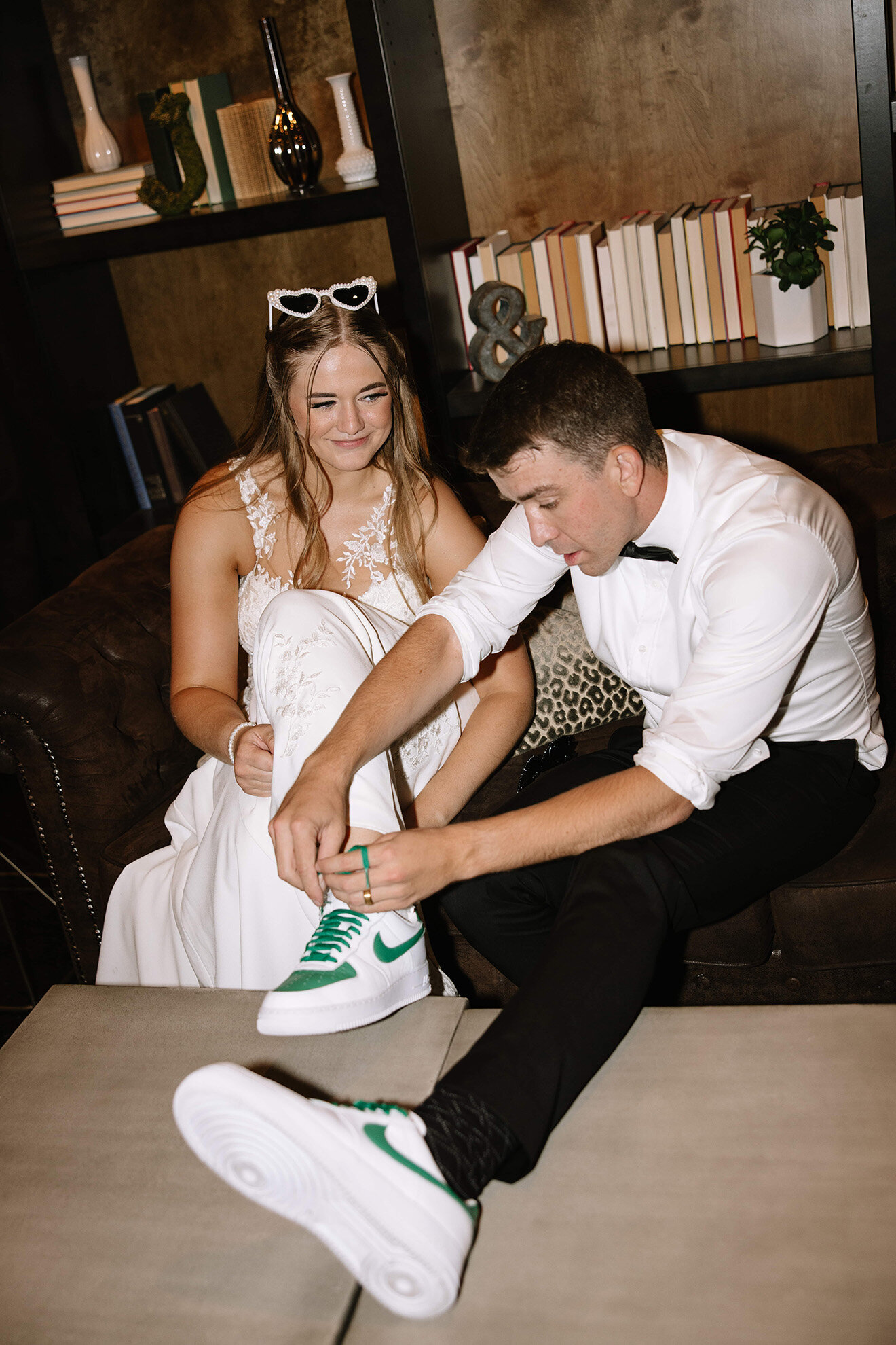 A groom laces up green Nike sneakers while sitting beside his smiling bride in a white gown at SoHo63, a Chandler wedding and event venue. The relaxed moment, set against a stylish lounge backdrop, captures the personality and charm of modern weddings at SoHo63.
