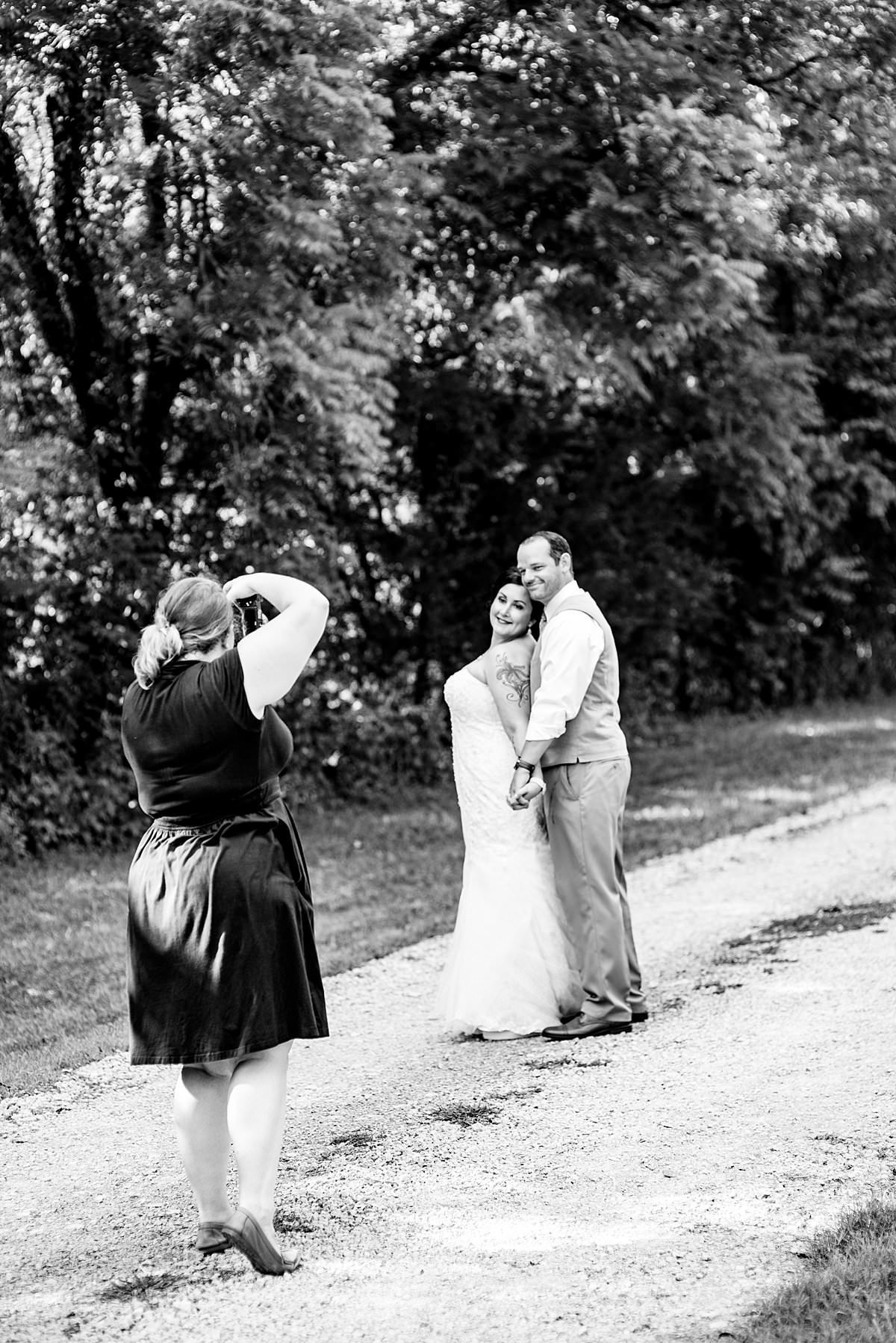 Black and White behind the scenes photo of owner taking photo of bride and groom on gravel road