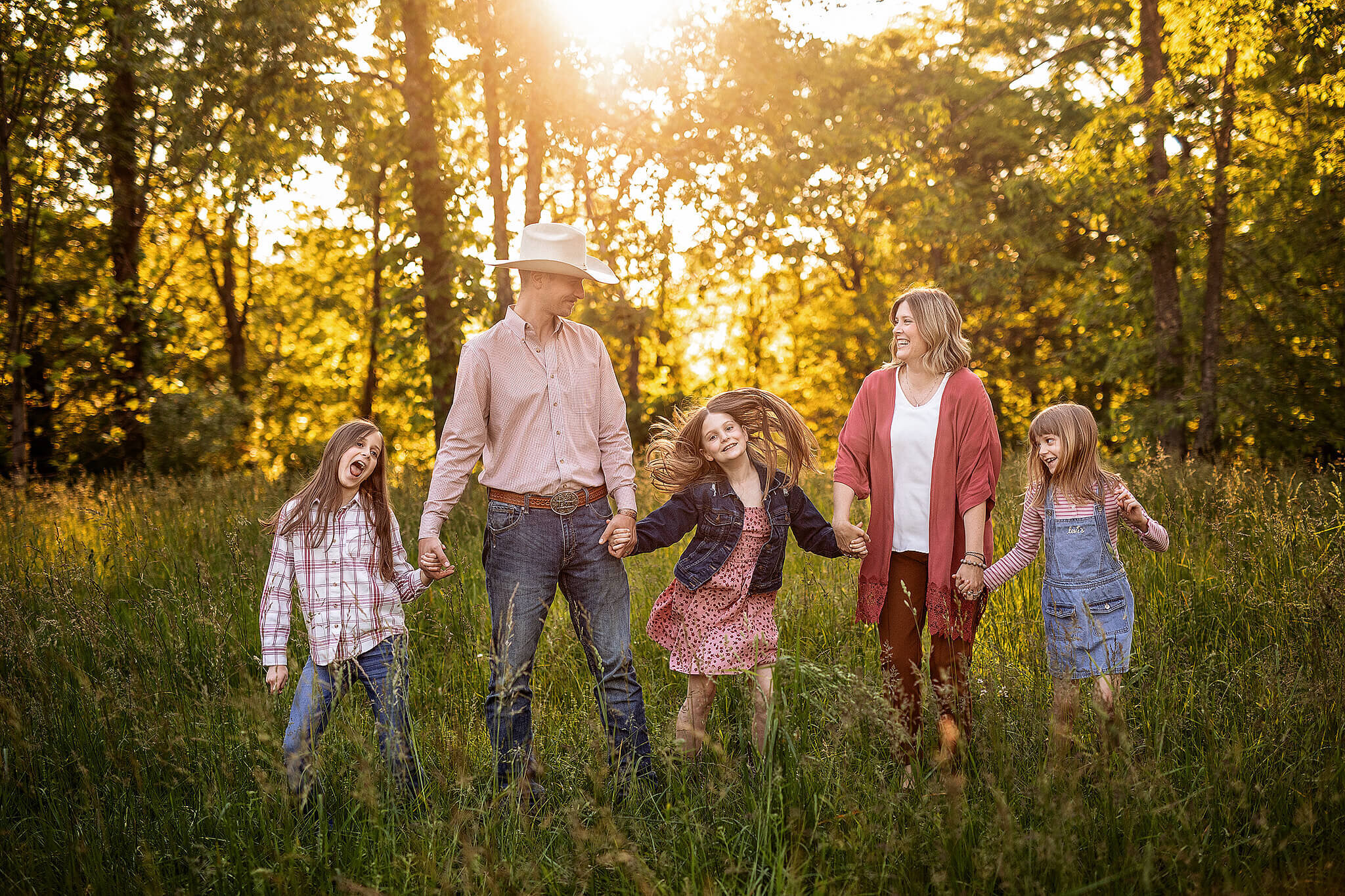 Family of 5 holding hands standing in a row in tall grass with the sun behind them