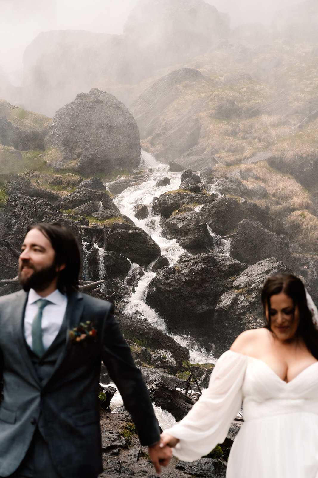 a couple in their wedding attire walk towards their adventure elopement photographers as they hold hands. The focus is on the waterfall behind them, using the couple to frame the landscape, offering a unique perspective