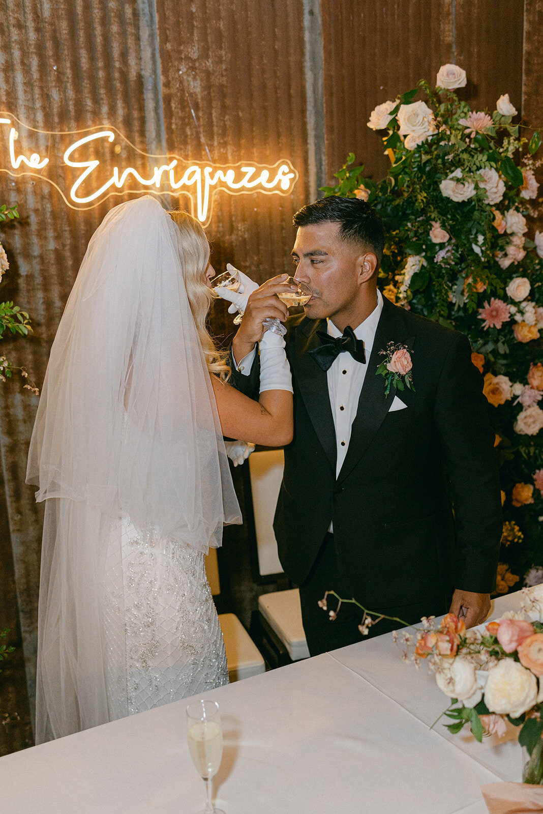 Bride and groom toasting with champagne in front of a custom neon last name sign surrounded by lush floral installations at their elegant Austin wedding reception