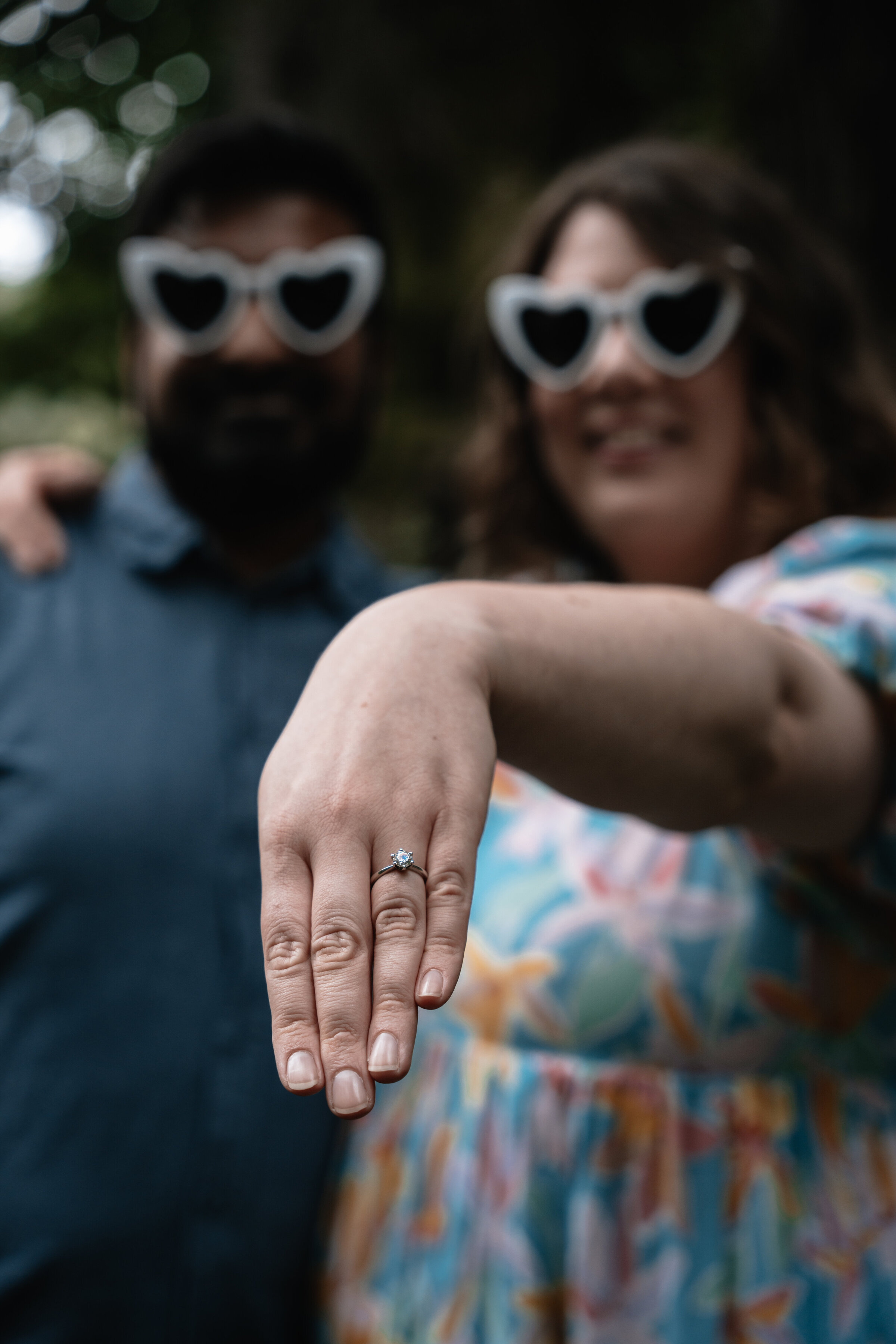 Close up of womans hand with engagement ring, in background couple are blurred but seen wearing heart shaped glasses