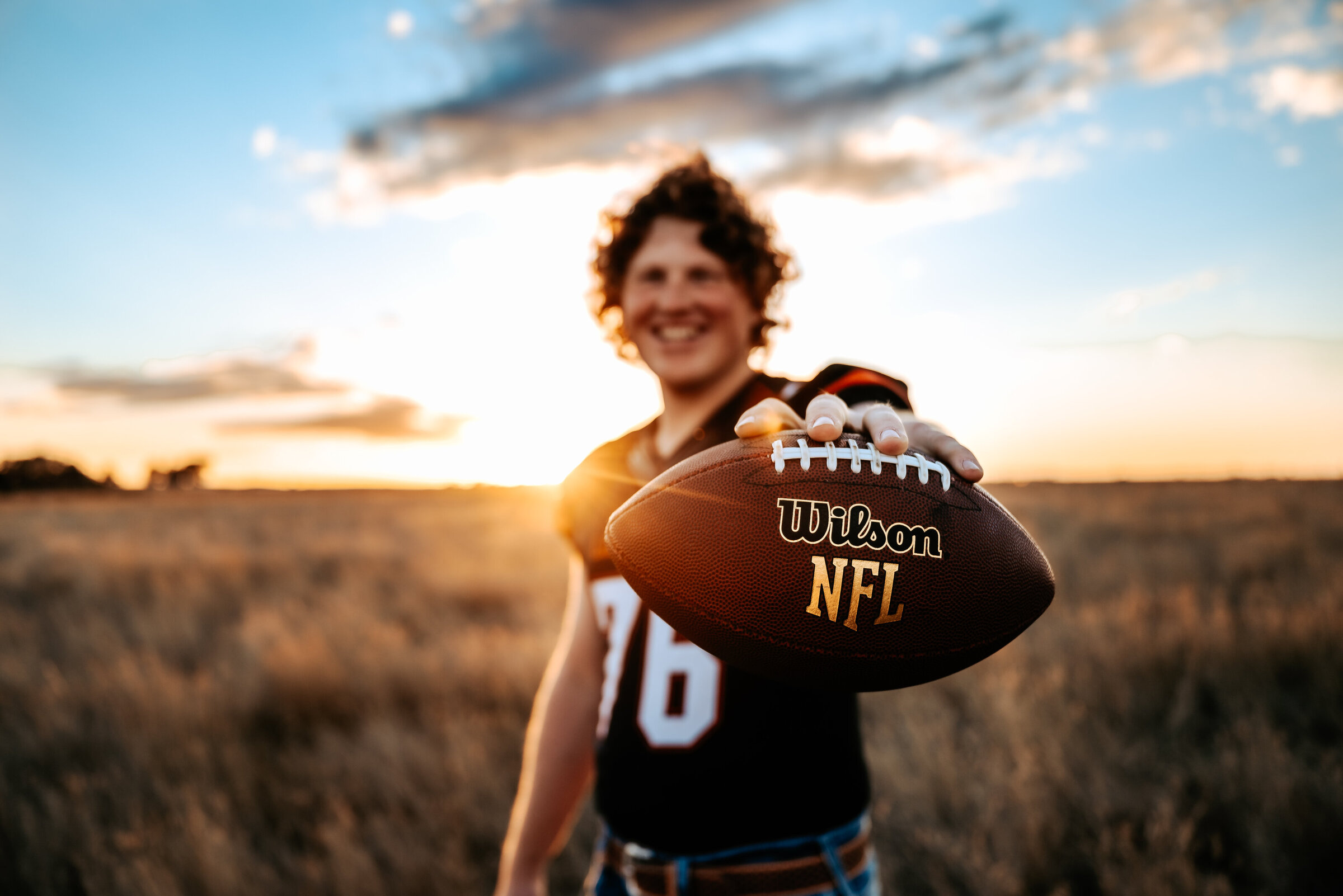 Senior wearing football jersey holds football in field at sunset