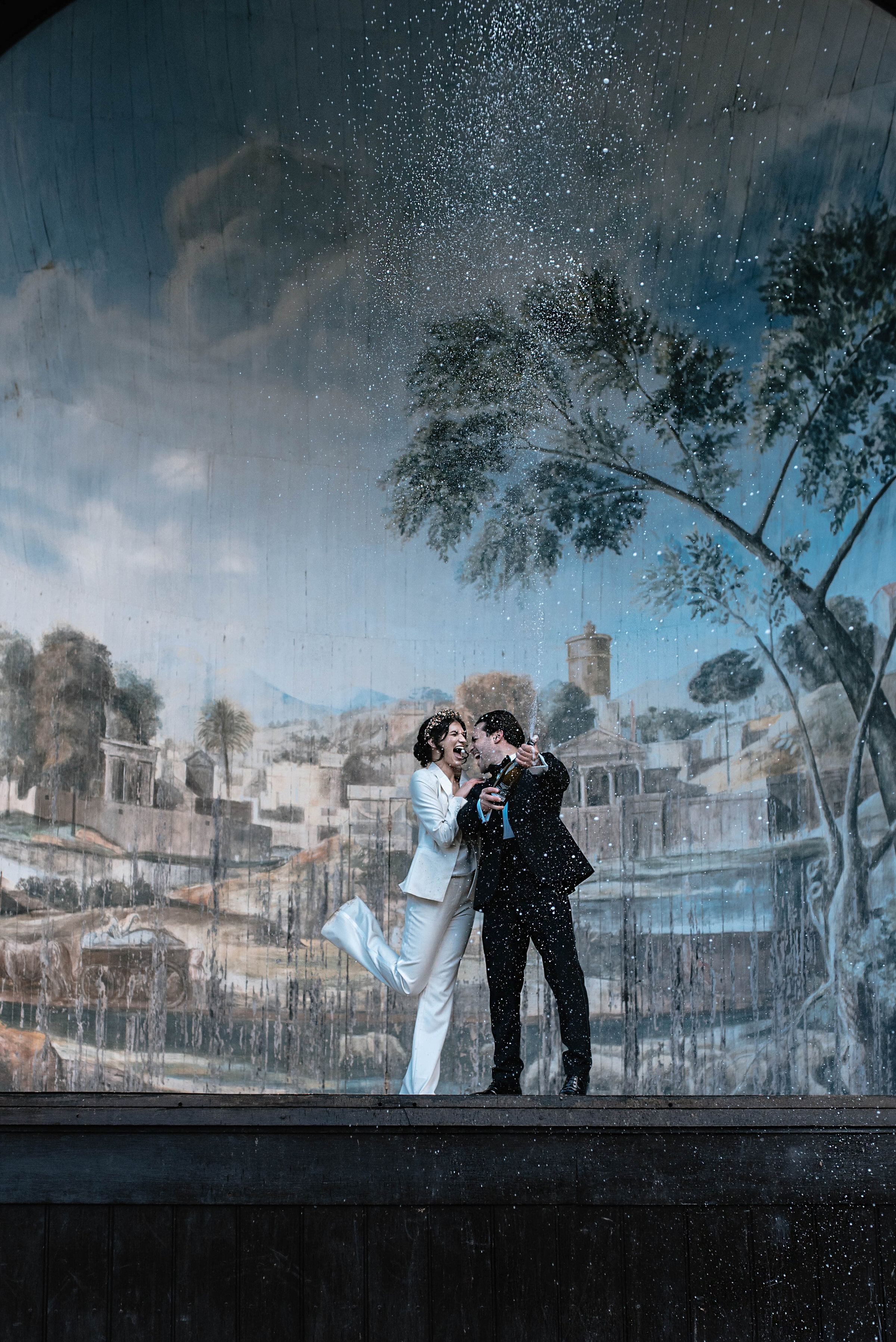 Bride and Groom spraying champagne in front of beautiful painted backdrop at Larmer Tree Gardens in Wiltshire