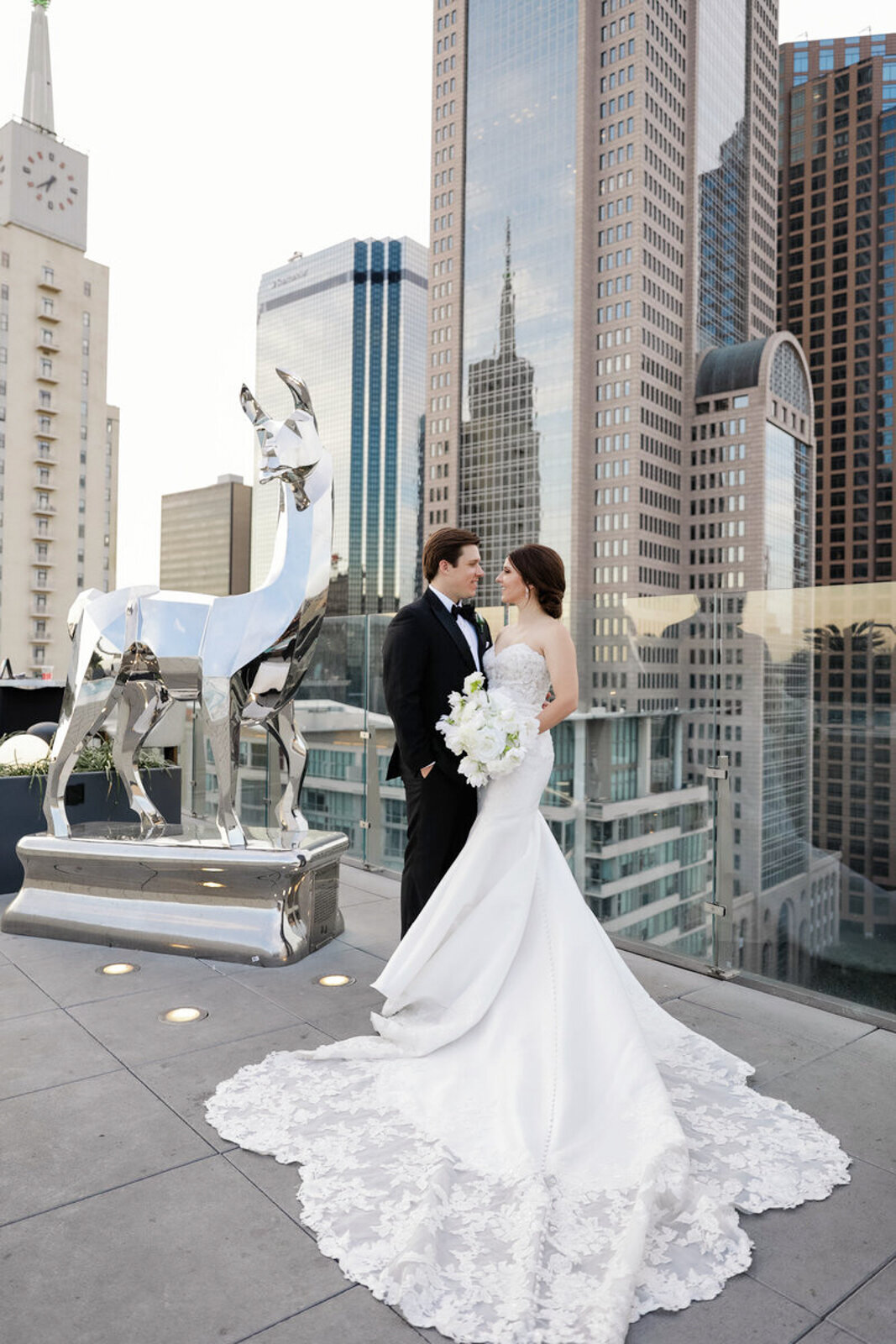 Bride and groom on the rooftop of The Statler in Dallas, standing beside a llama statue with the city skyline in the background.