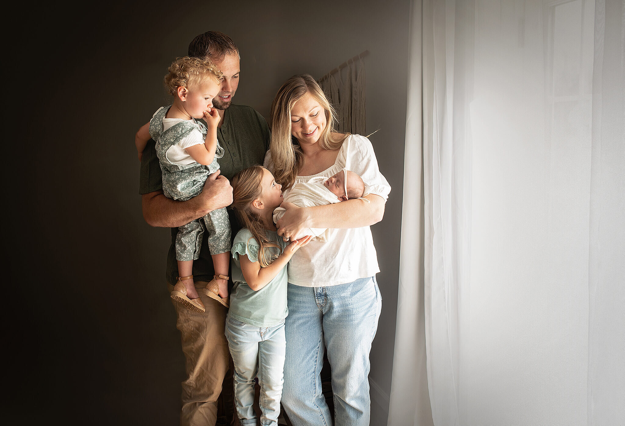 Family of 5 with their newest baby standing next to a window looking at their newest addition