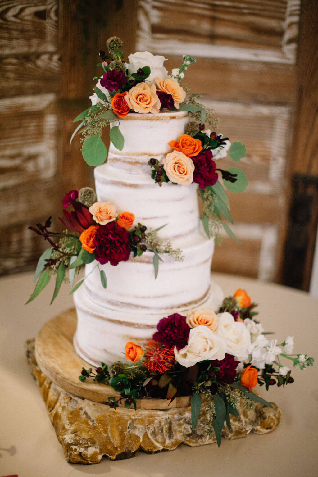 A three-tiered wedding cake decorated with orange, red, and white flowers, green foliage, and set on a wooden slab against a wooden background.