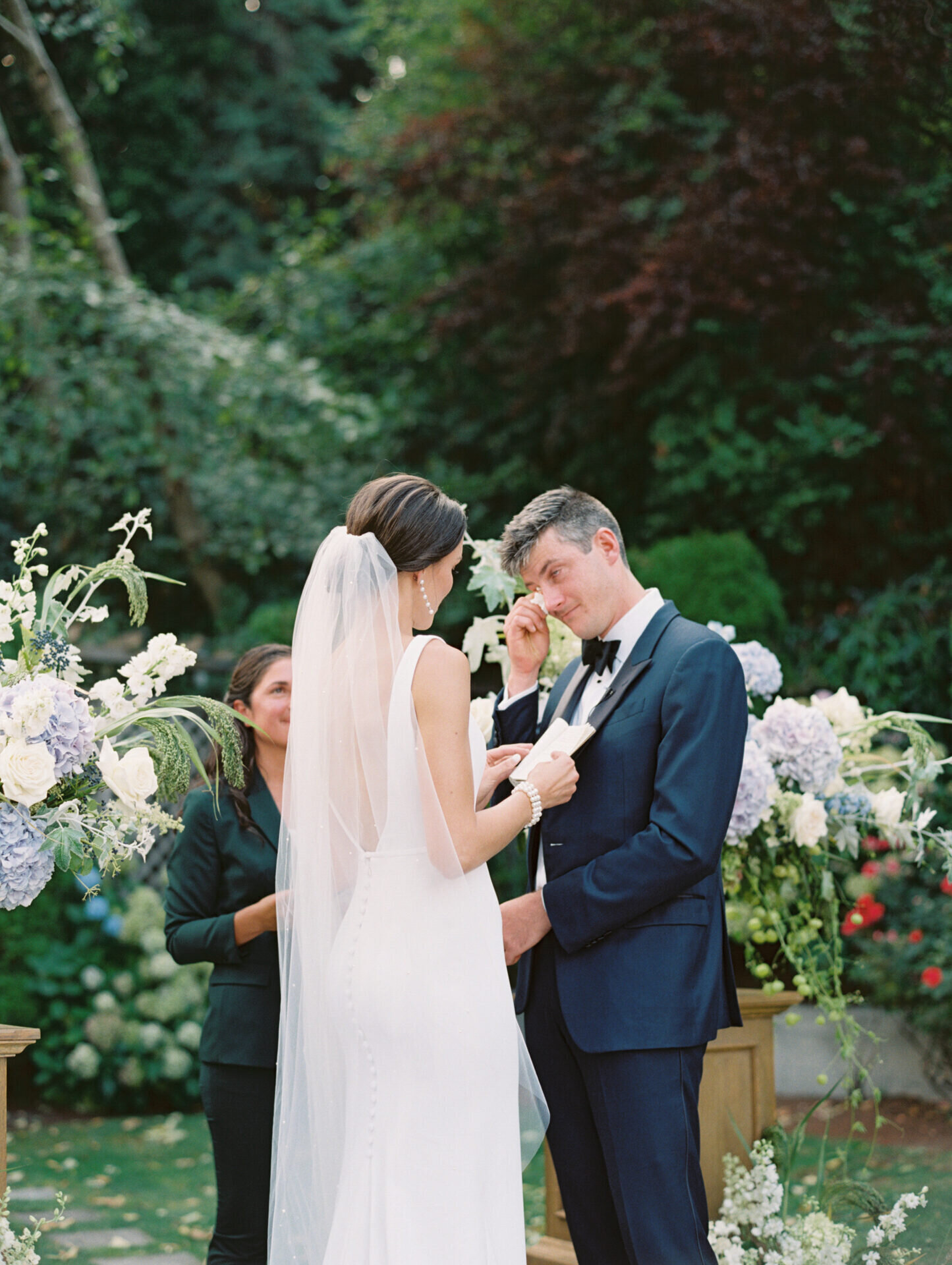 groom in navy suit crying at wedding ceremony with greenery background