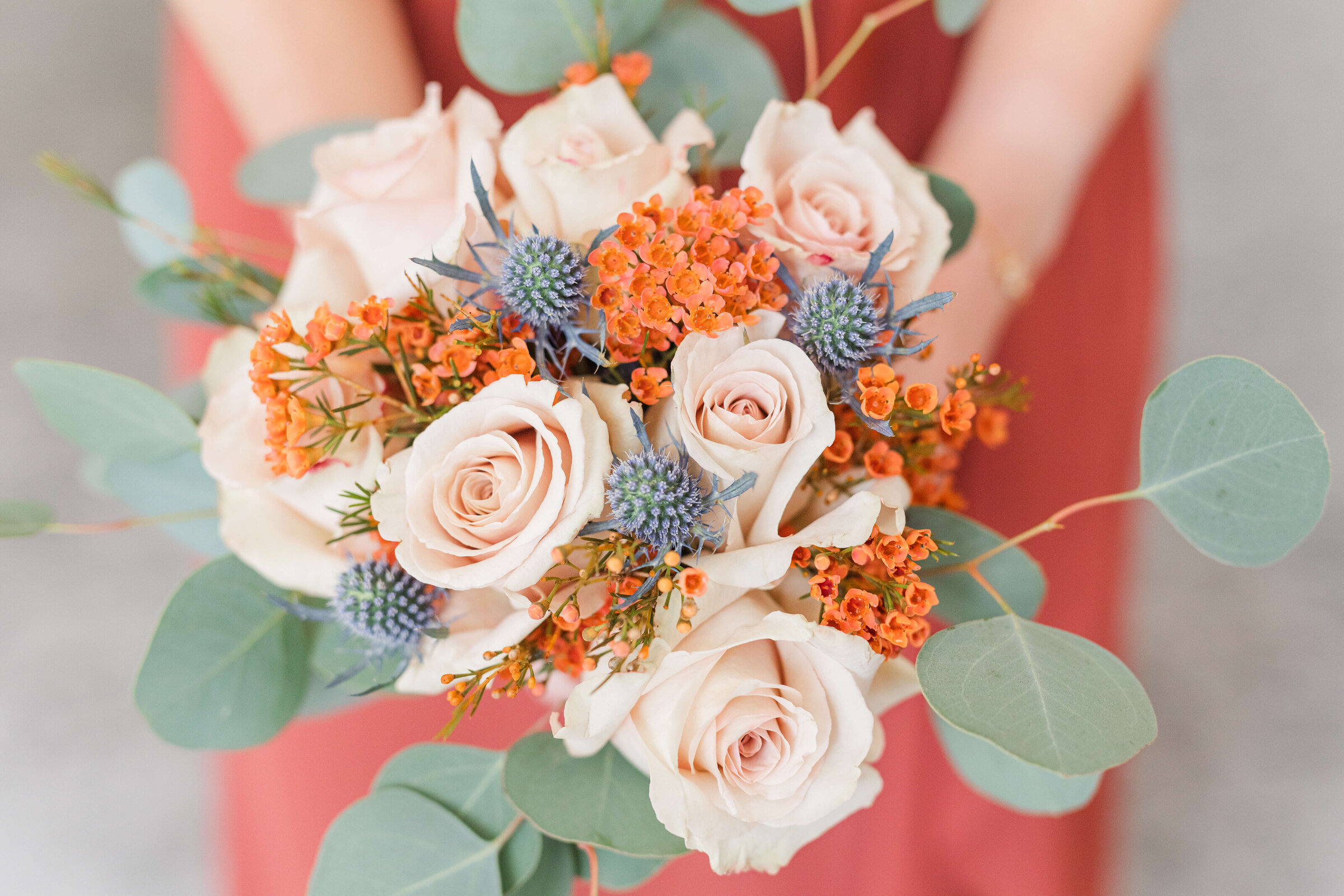 an up close photo of a wedding bouquet that has peach, navy, and orange flowers in it with greenery. The bouquet is held by a bridesmaid in an orange dress.