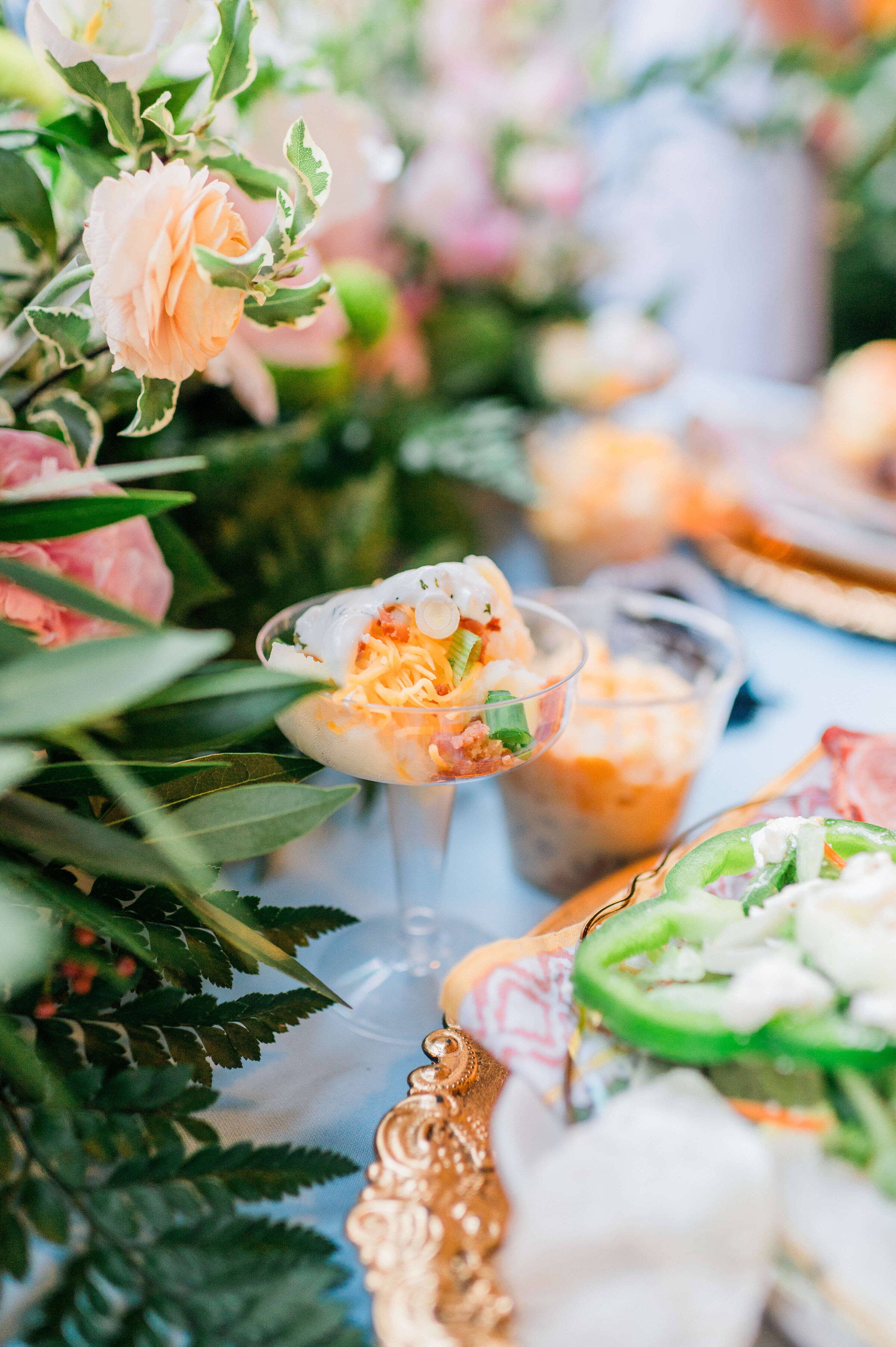 A close-up of a festive table setting with a glass filled with a creamy shrimp appetizer, garnished with herbs and cheese, surrounded by lush greenery and pastel flowers.