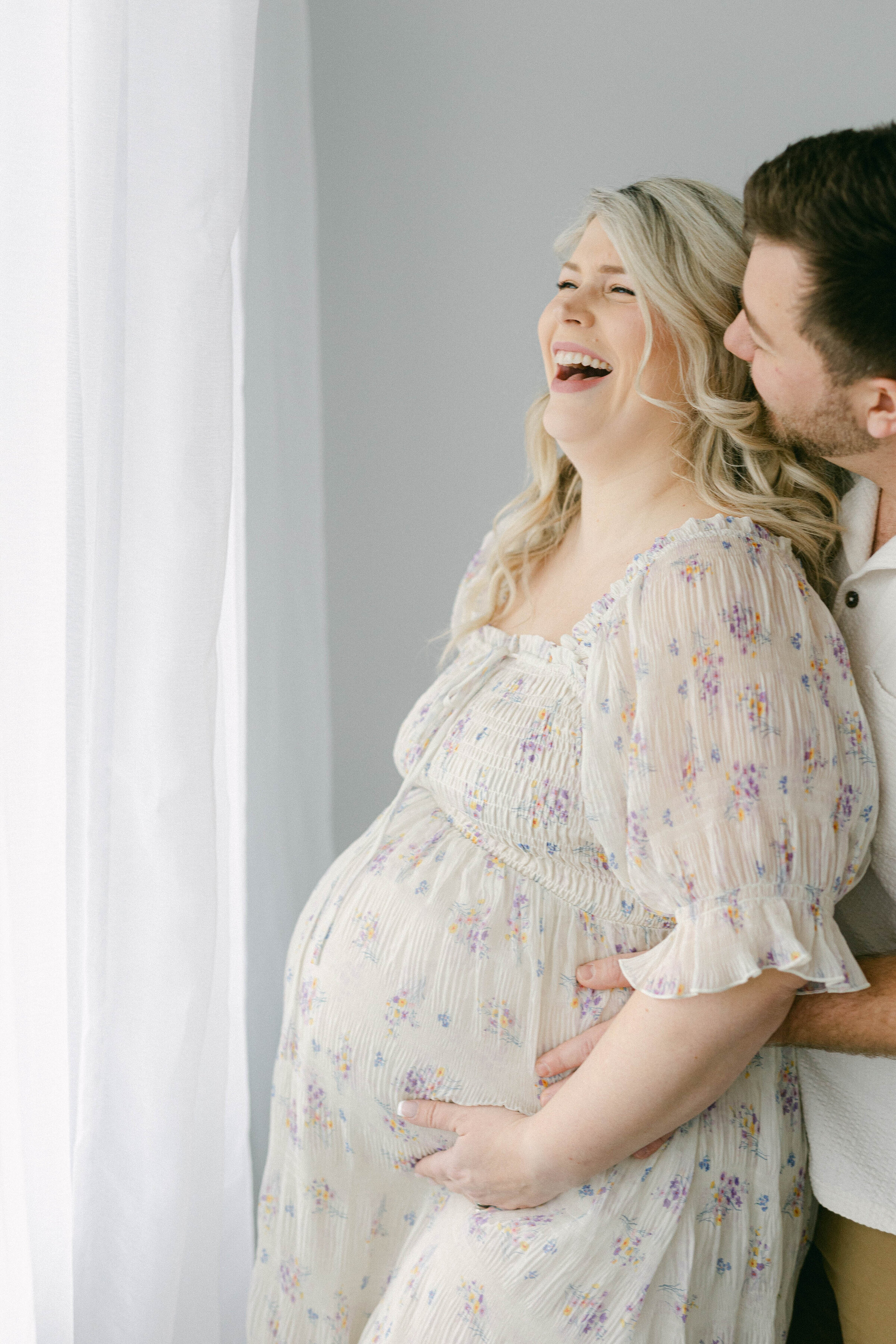 man wrapping arms around womens pregannat belly while she smiles big looking out the window  knoxville tennessee newborn photographer