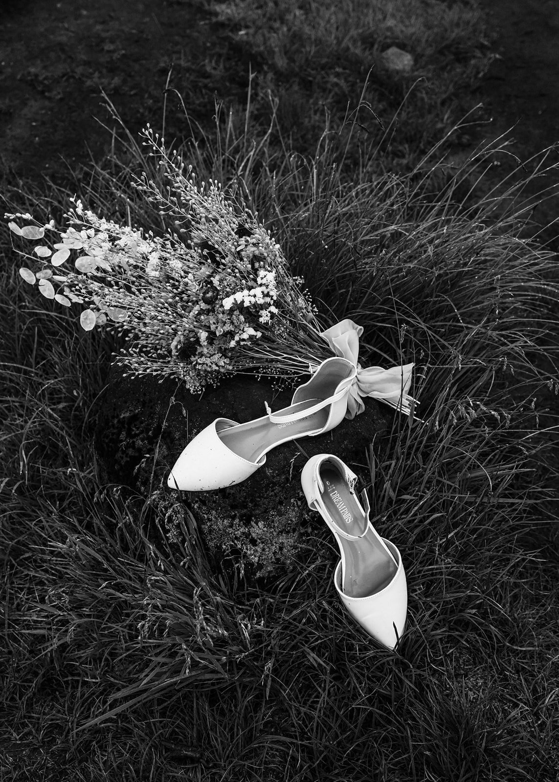a black and white image of details from tan adventure elopement. we see a pair of white heels and a bouquet laying in the tall grass.