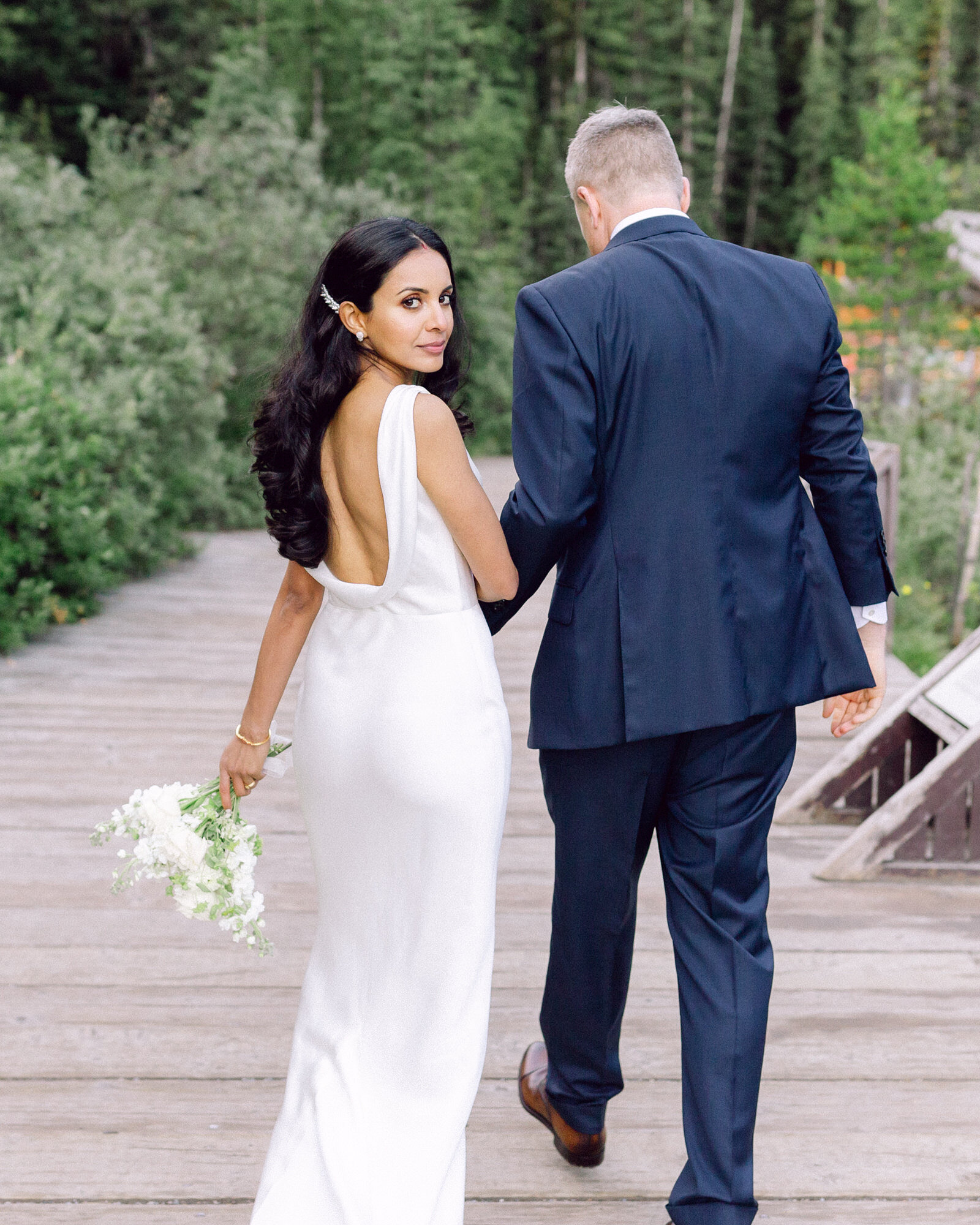 A stunning elopement photograph capturing a bride as she looks back at the camera while holding hands with her husband near Chateau Lake Louise in Banff National Park. As they walk toward the iconic boathouse, this moment embodies elegance, romance, and the breathtaking beauty of their intimate wedding day.