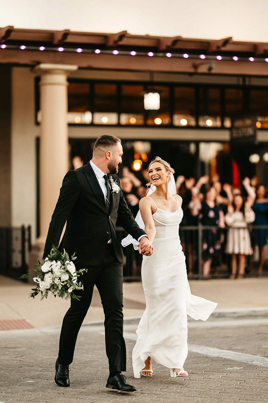 A bride and groom walk hand-in-hand past cheering guests outside SoHo63, a Chandler wedding and event venue. The bride looks up at her groom with a joyful smile while holding a bouquet, capturing the excitement and charm of a downtown Chandler wedding celebration.