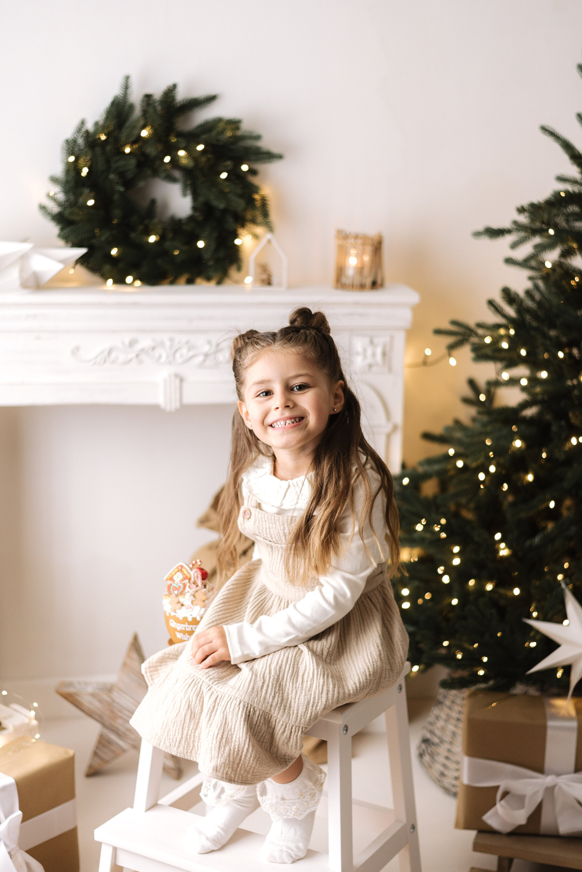 Little girl smiling sitting on a white seat infront of christmas tree and fairy lights at billingshurst christmas photoshoot