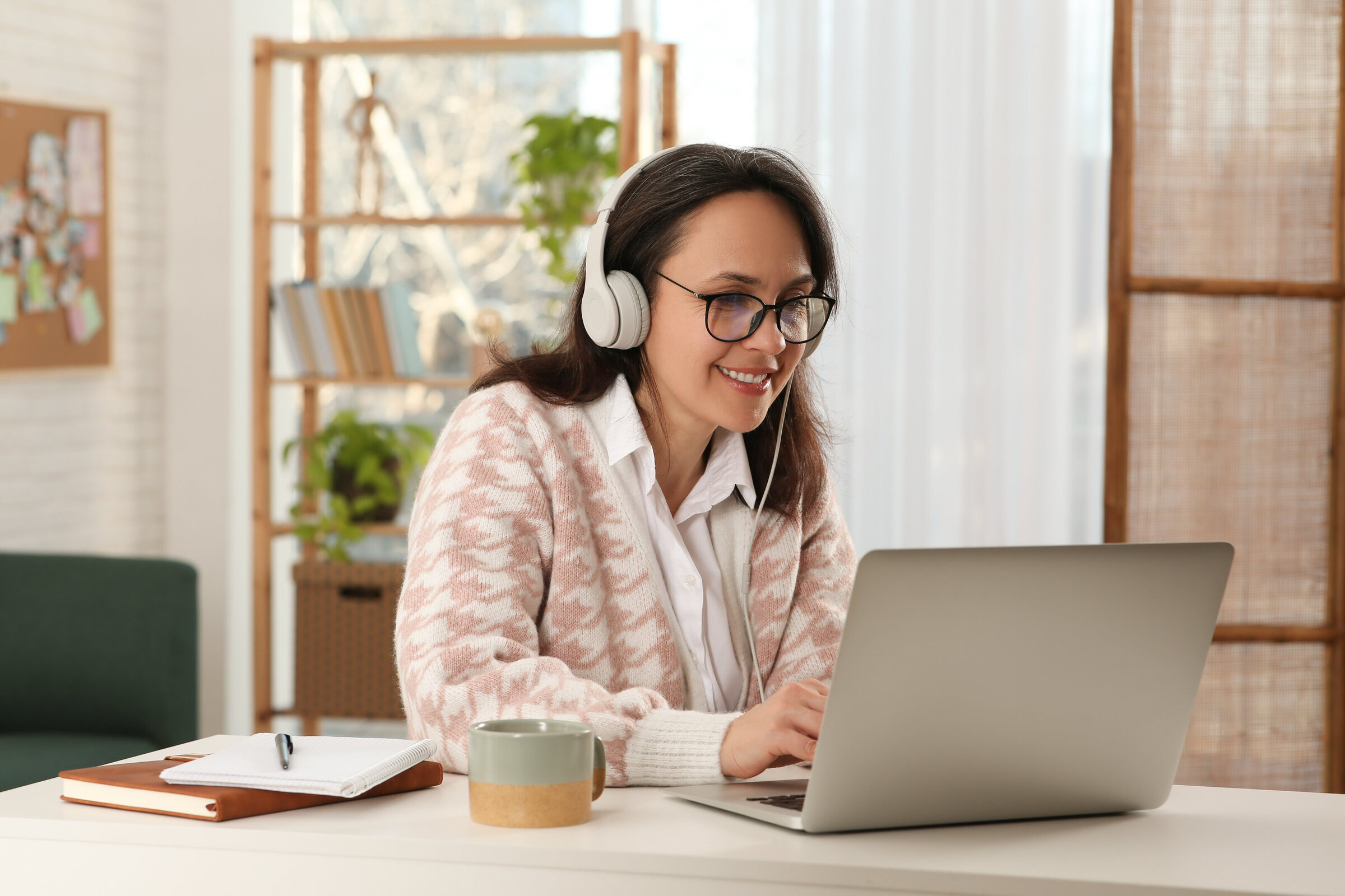 A smiling woman with headphones working at a laptop in a warm, plant-filled home office