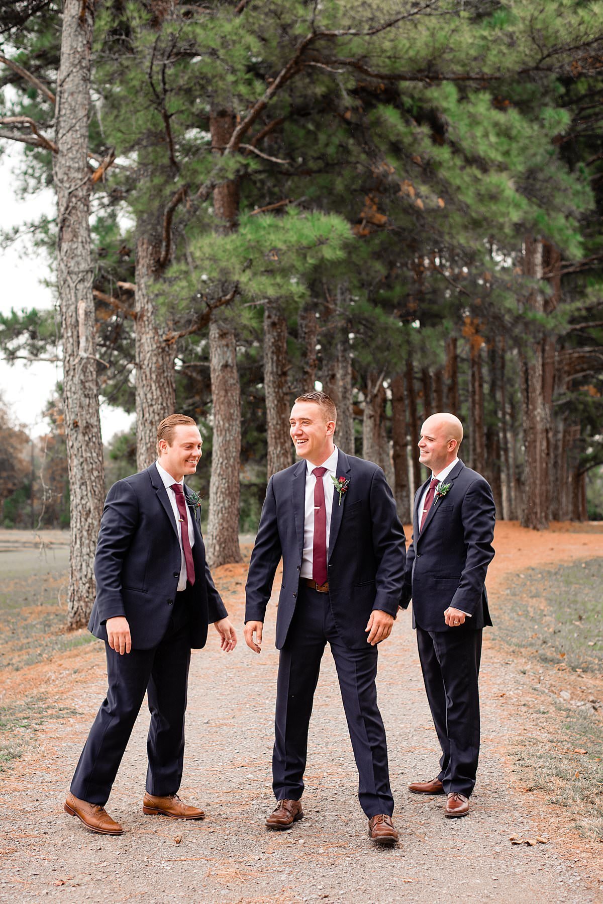 Groom and his 2 groomsmen laughing while on a gravel path with tall evergreen trees behind them