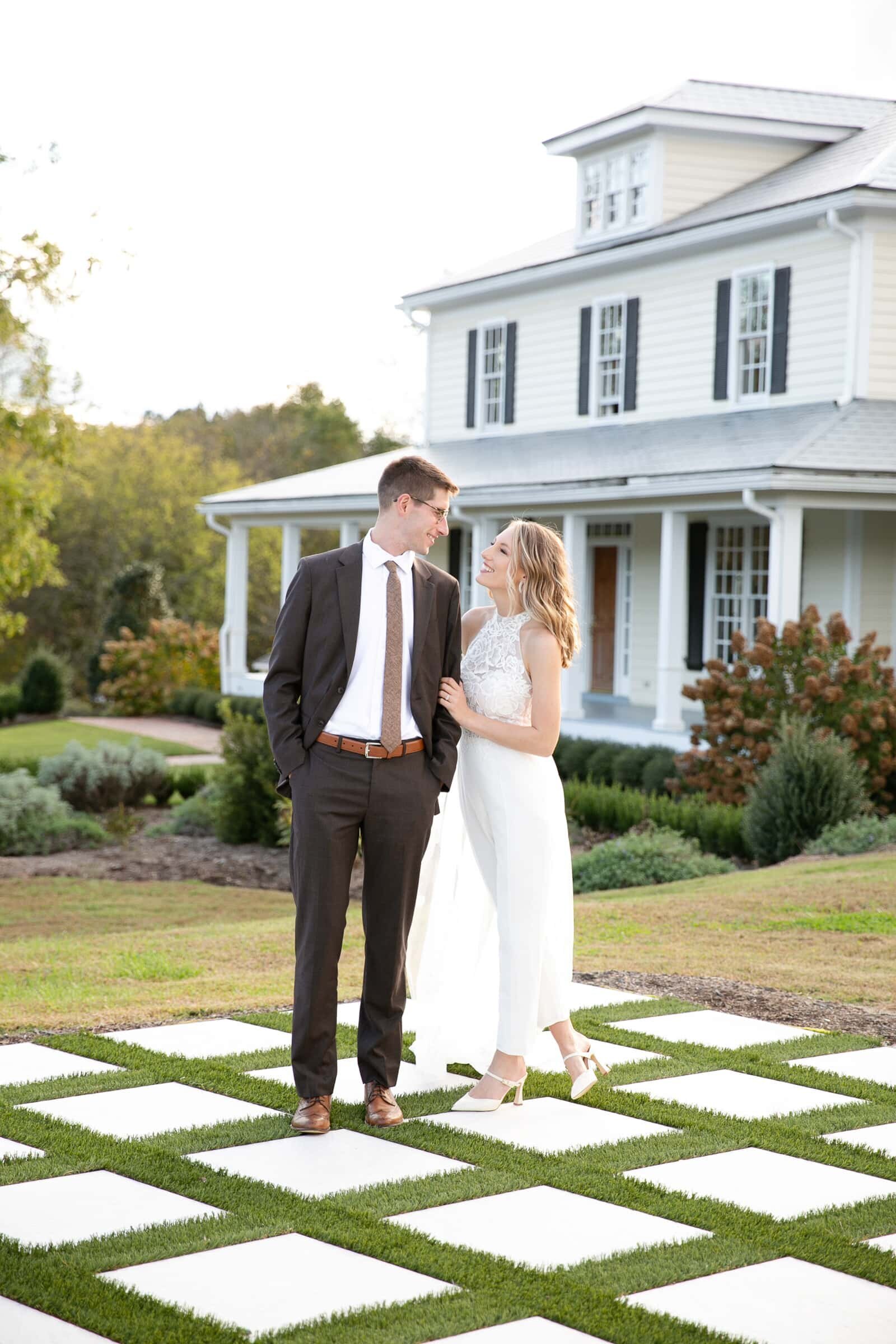 Wedding couple looking into each other's eyes standing in front of the main house at Walnut Hill Raleigh. The woman is wearing a white pantsuit and the man is wearing brown suit.