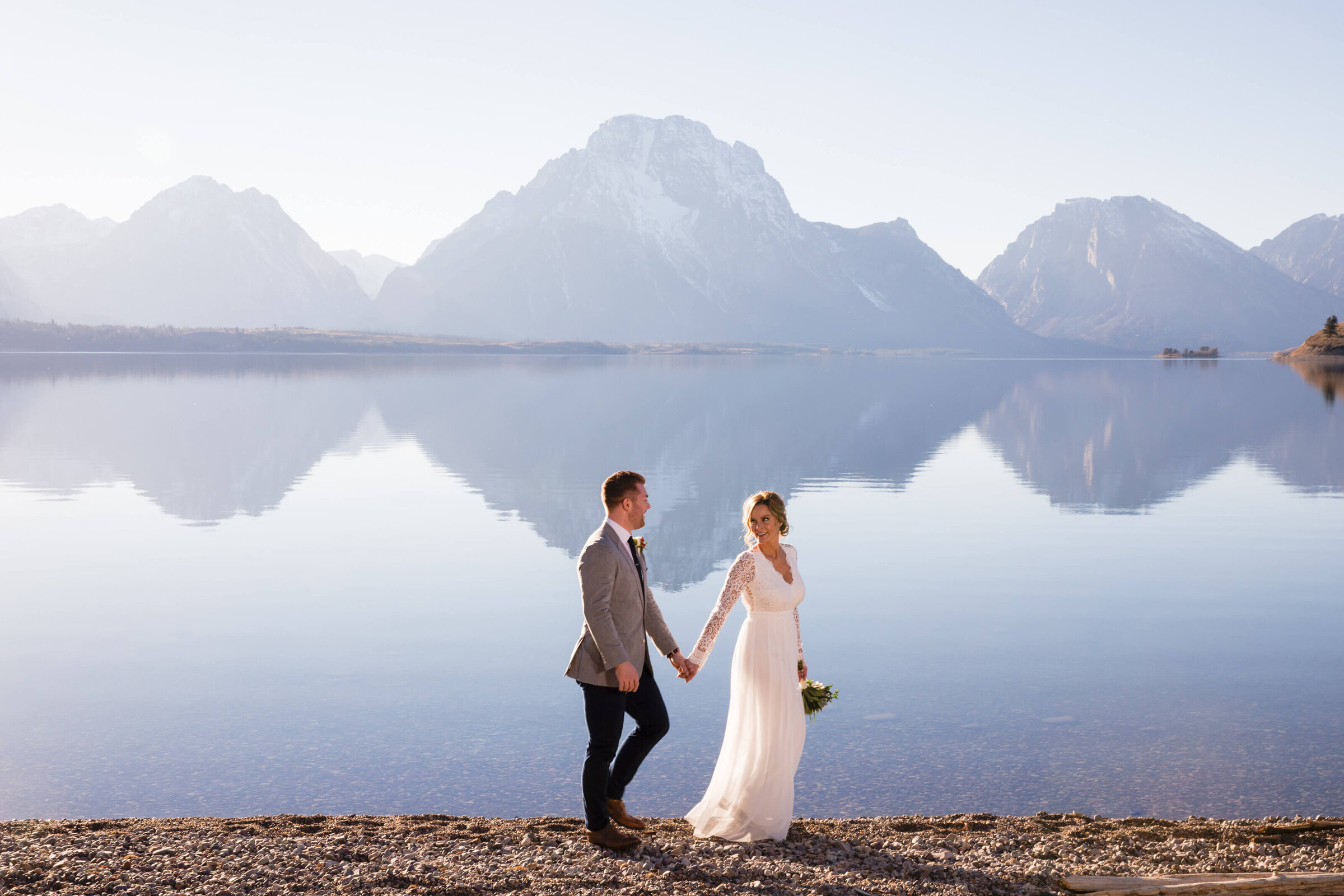 A newlywed couple walks hand in hand along the shore of Jackson Lake, surrounded by the Teton Range reflecting in the still water. The bride wears a lace-sleeved wedding dress and carries a bouquet, while the groom, dressed in a light gray suit, gazes at her. A serene and romantic wedding moment in Grand Teton National Park.