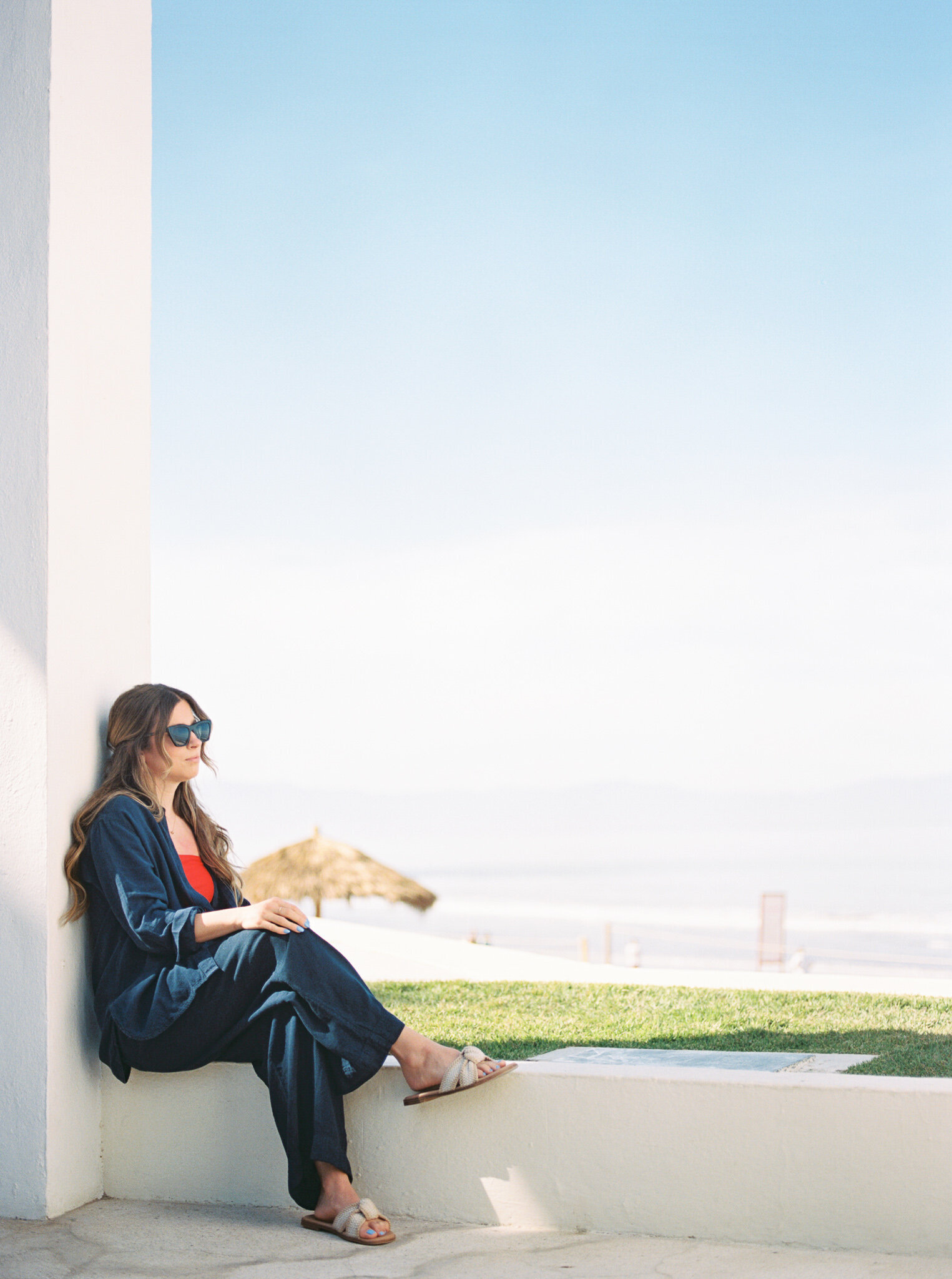 Photographer Little Rock sits on a white building ledge at the beach in a blue and red outfit, looking out towards the water.