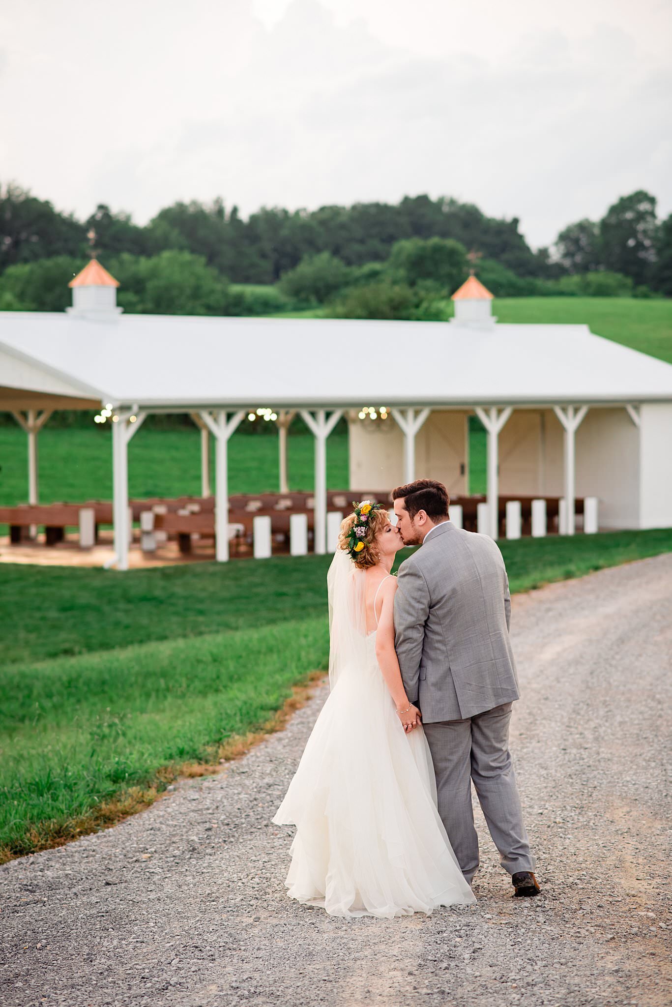 Bride and Groom walking hand in hand down gravel road towards the ceremony pavilion at White Dove Barn, they paused to lean in for a kiss
