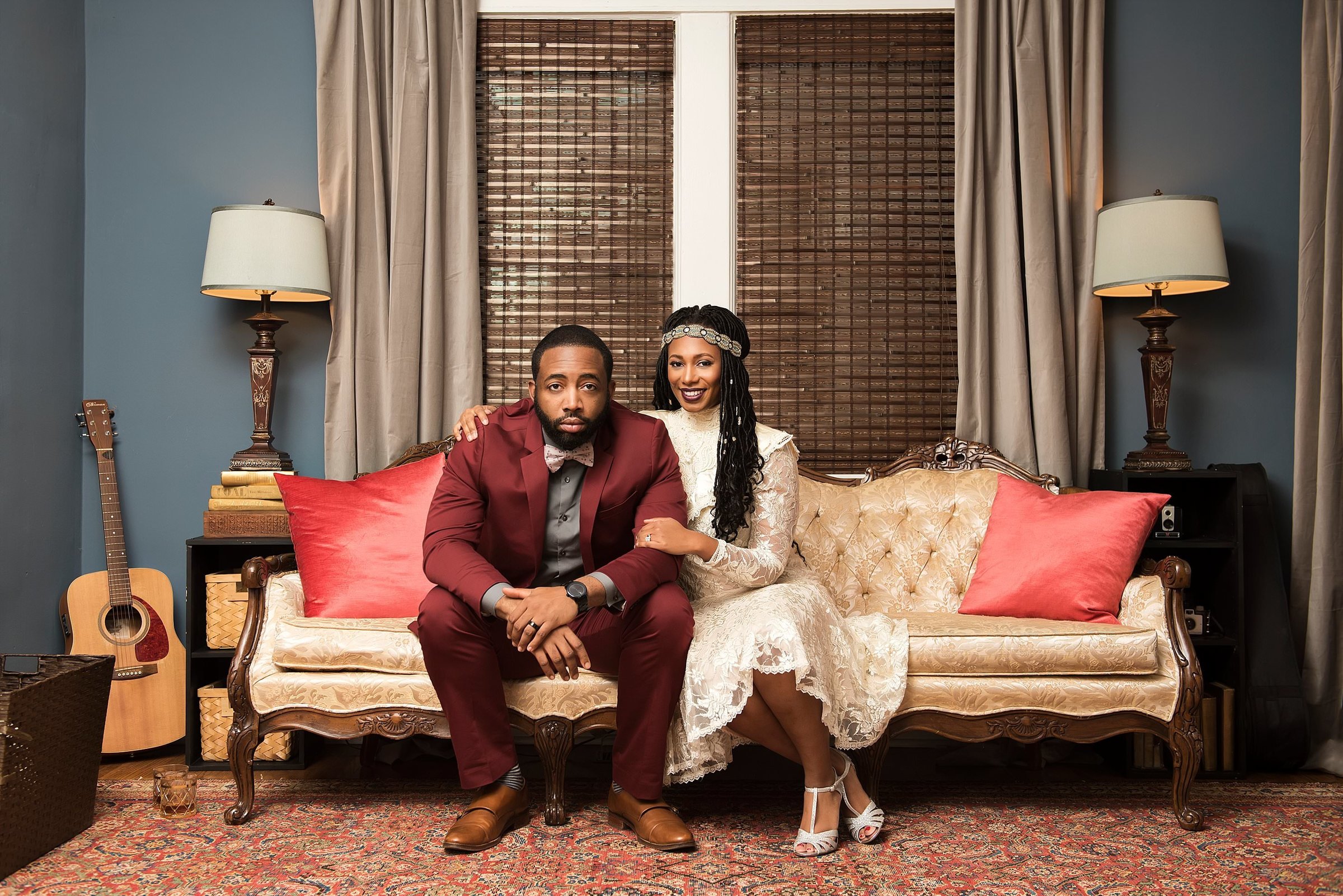 Couple sitting on a vintage couch in living room, he is wearing a retro maroon suit and she has a vintage wedding dress on