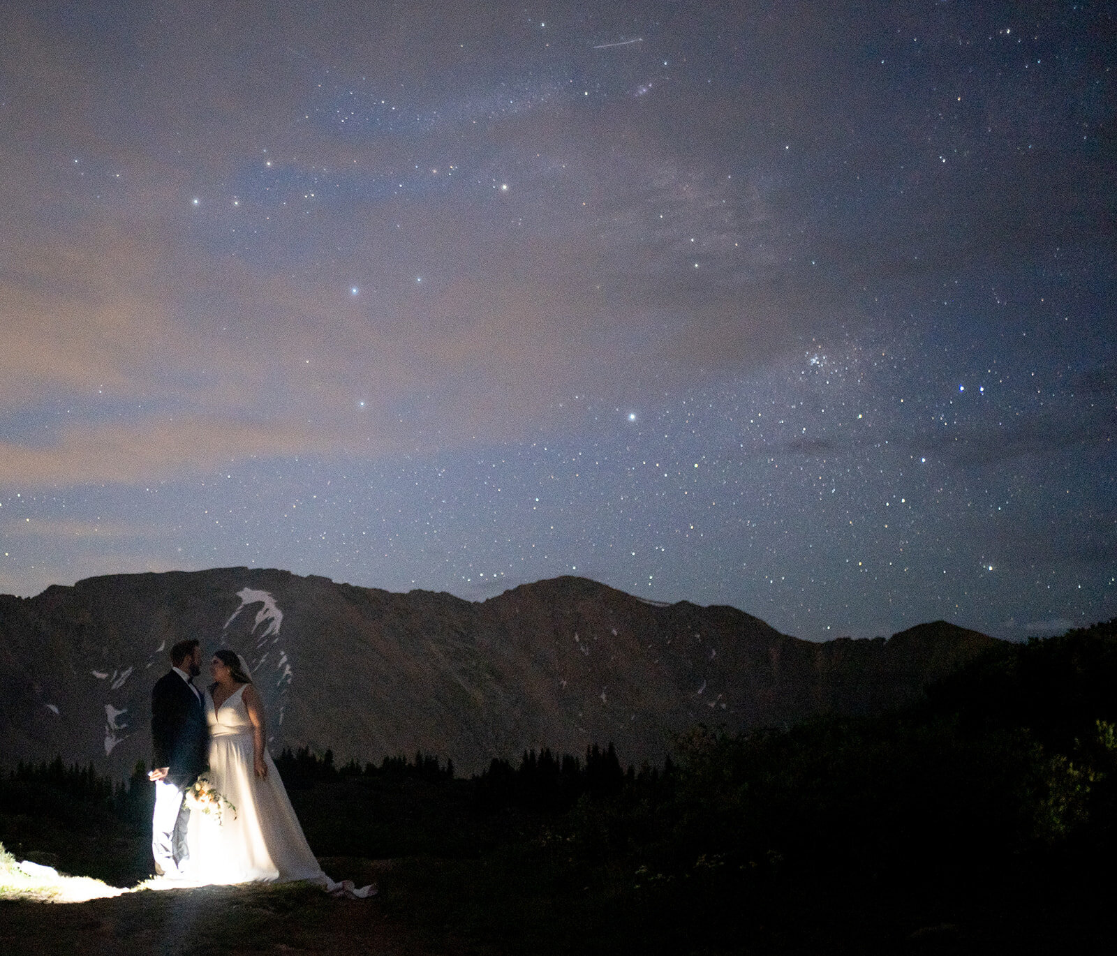 bride and groom are standing beneath the stars holding lanterns. they are looking at each other and smiling and there is a purple/blue sky with mountains in the background.